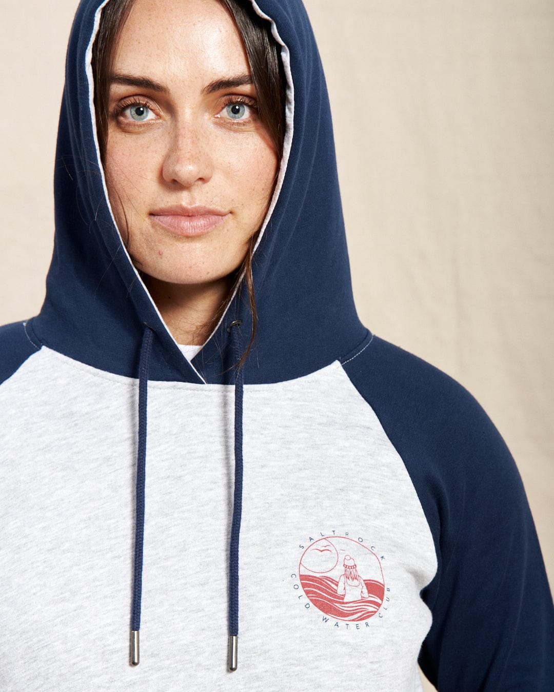 A person wears the Saltrock Coldwater Club Women's Raglan Pop Hoodie in navy and gray, featuring contrasting sleeves and a circular logo on the chest with a wave design and "Antarctic Coldwater" text.