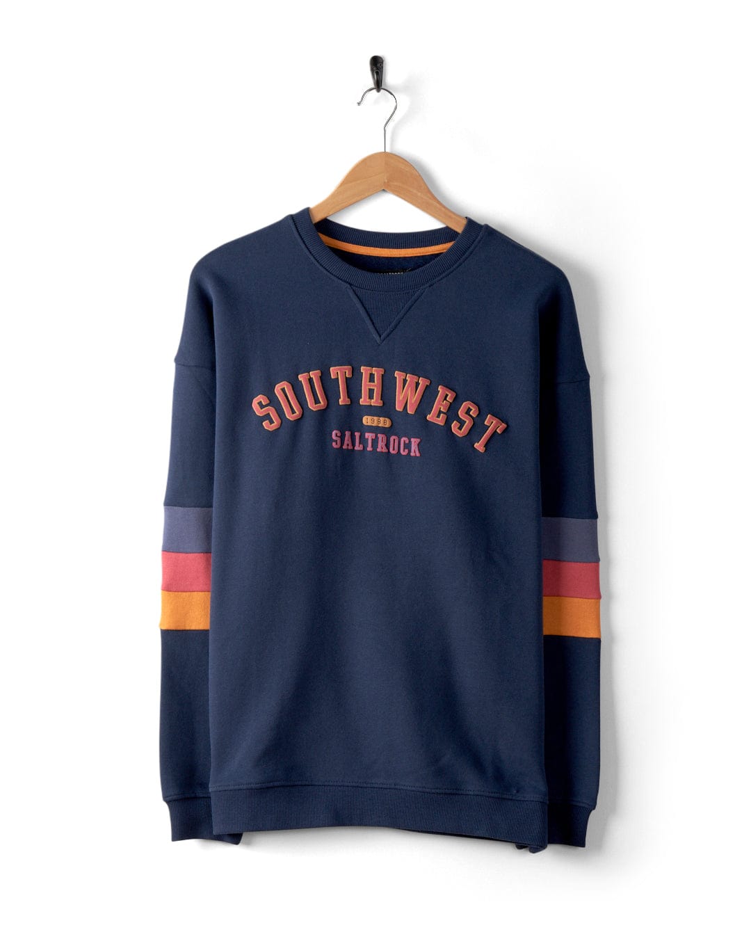 A navy blue sweatshirt from Saltrock, named "College - Womens Oversized Sweat - Blue," hangs on a wooden hanger with "SOUTHWEST SALTROCK" printed in vibrant orange and yellow text. The sleeves feature charming retro stripes in red, orange, and purple. This oversized fit is a stylish homage to the Southwest 1988 aesthetic.