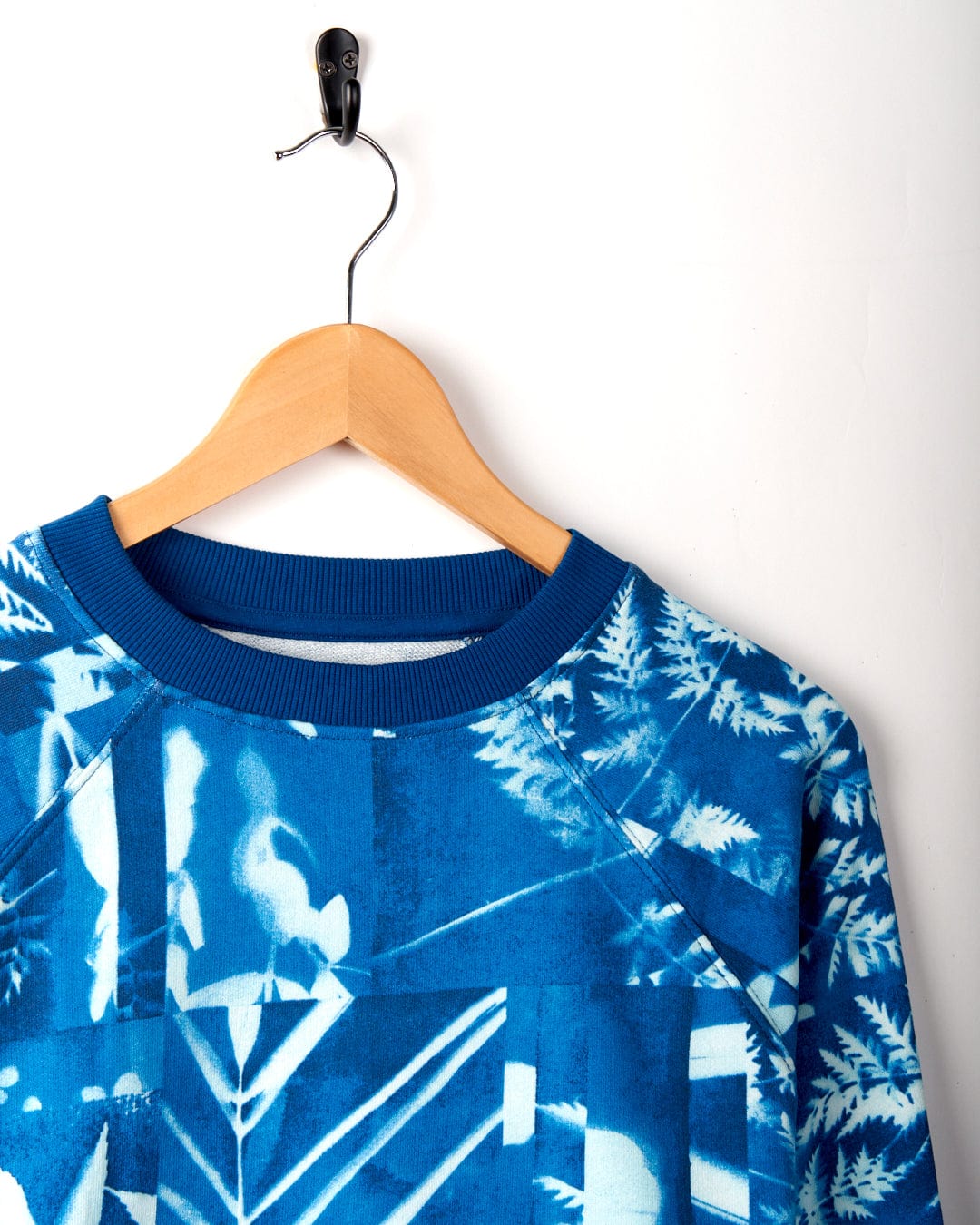 A Saltrock Cyanotype - Womens Recycled Boxy Sweat - Blue hangs on a wooden hanger against a white wall, suspended by a black hook.