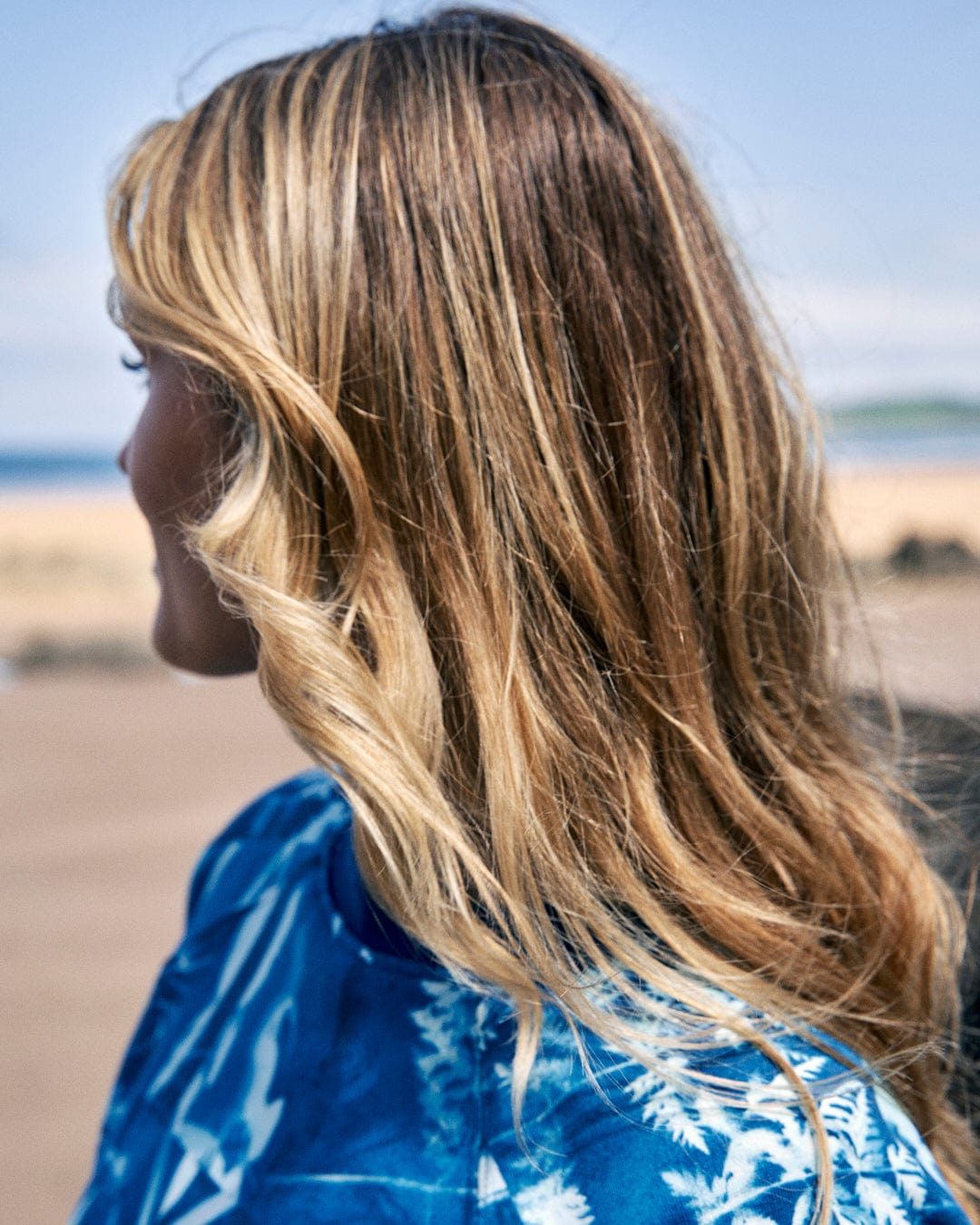 A person with long, wavy blonde hair is facing away from the camera, standing on a sandy beach and wearing a Cyanotype - Womens Recycled Boxy Sweat - Blue by Saltrock.