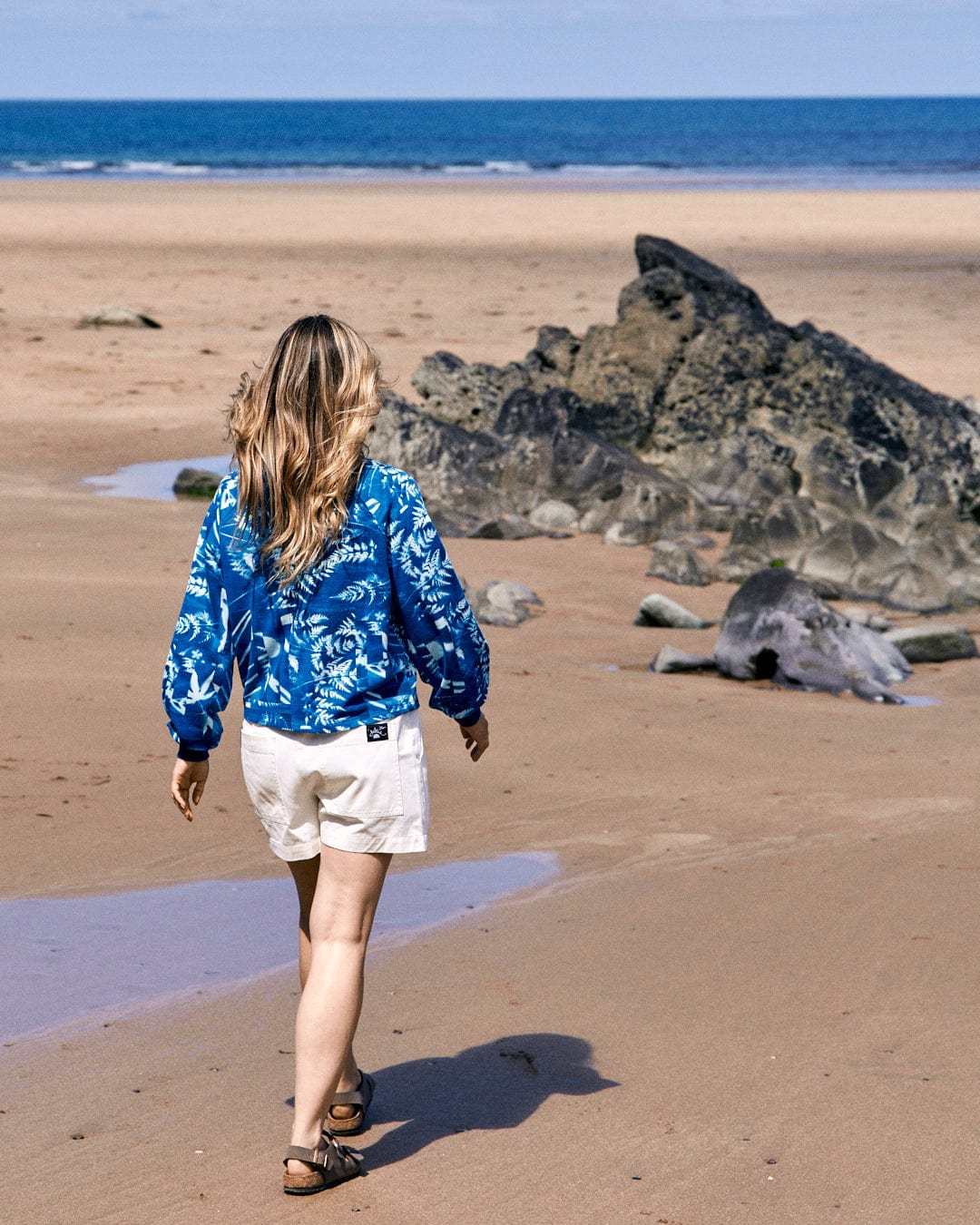 A person wearing a Cyanotype - Womens Recycled Boxy Sweat - Blue by Saltrock and white shorts with an abstract forest print walks along a sandy beach towards a rock formation.
