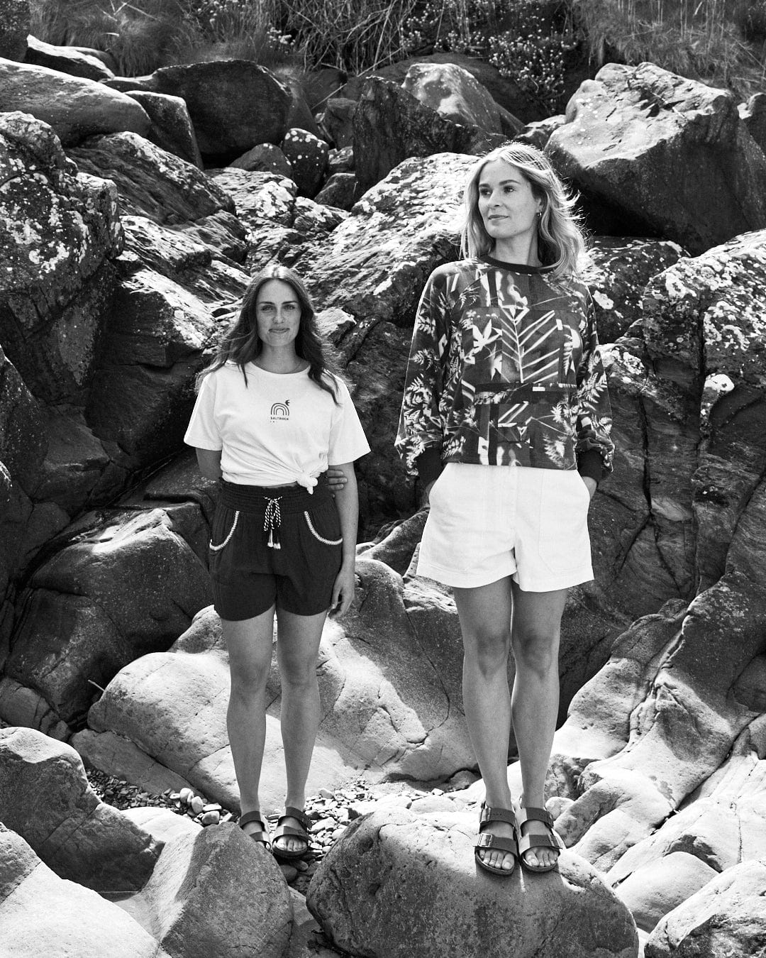 Two women stand among large rocks. One is wearing a Saltrock Cyanotype - Womens Recycled Boxy Sweat - Blue and shorts, while the other sports a patterned long-sleeve shirt featuring an abstract forest print and shorts. The background consists of rocks and some vegetation.