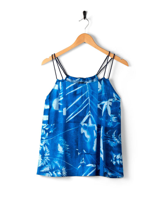 A blue sleeveless blouse with an abstract floral print and thin shoulder straps hangs on a wooden hanger, its lightweight material softly draping against the white background.