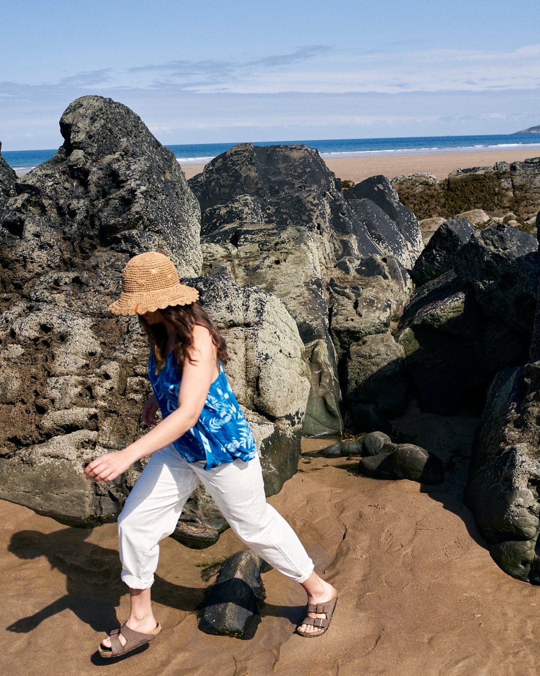 A person in a Saltrock Cyanotype - Womens Cami - Blue, white pants, and a straw hat walks among large rocks on a sandy beach with a view of the ocean in the background.