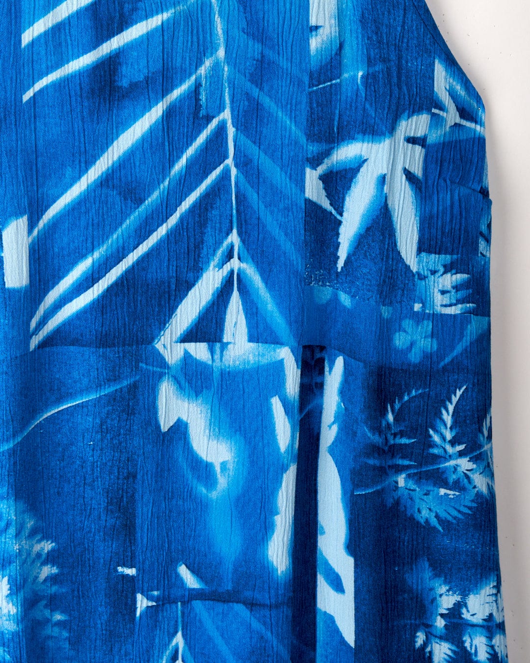 Close-up of a blue, crinkle viscose fabric featuring a white tropical leaf pattern and textured lines from the Cyanotype - Womens Cami - Blue by Saltrock.
