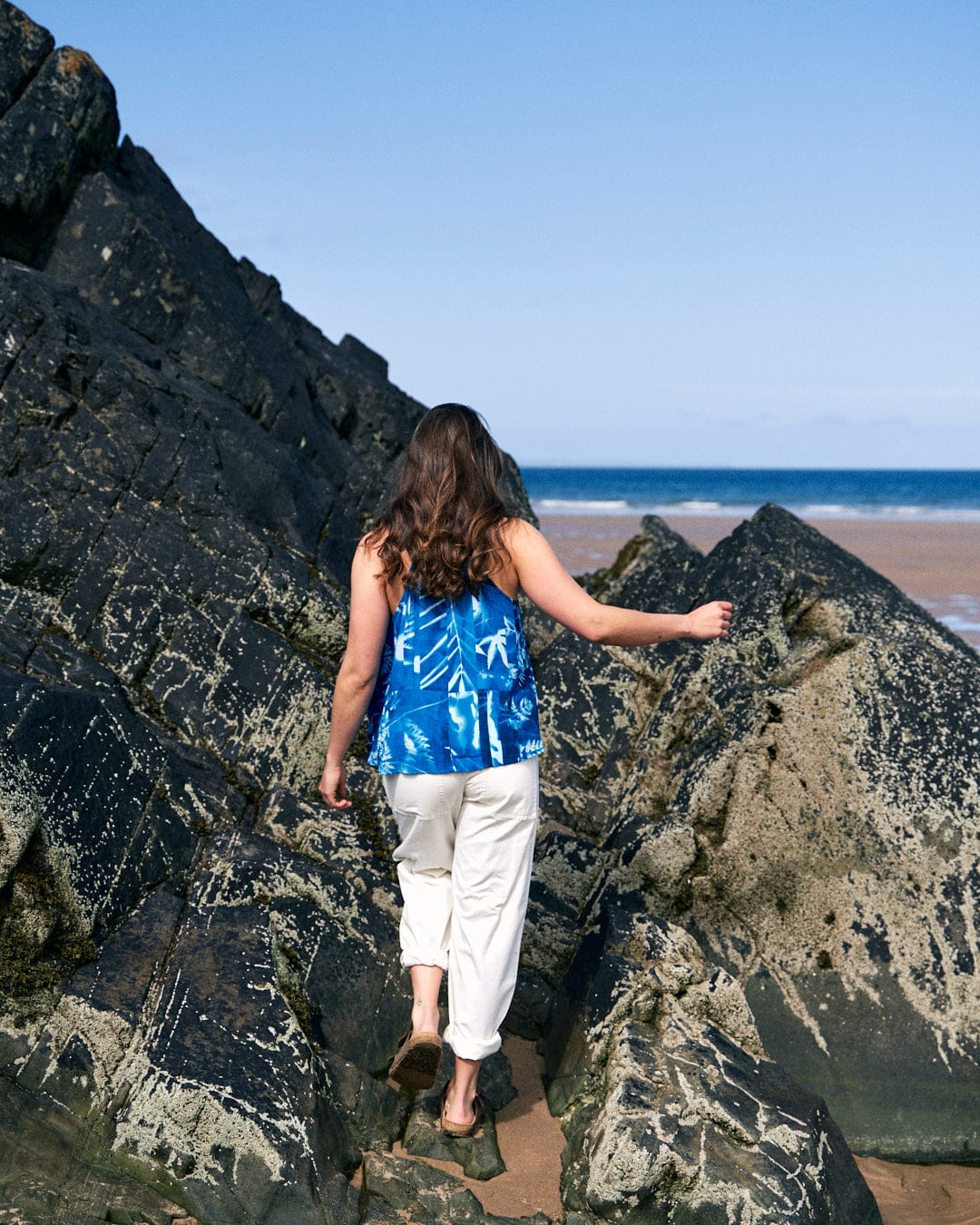 A person in a Saltrock Cyanotype - Womens Cami - Blue and white pants made of lightweight material walks on uneven, rocky terrain near a beach on a clear day.