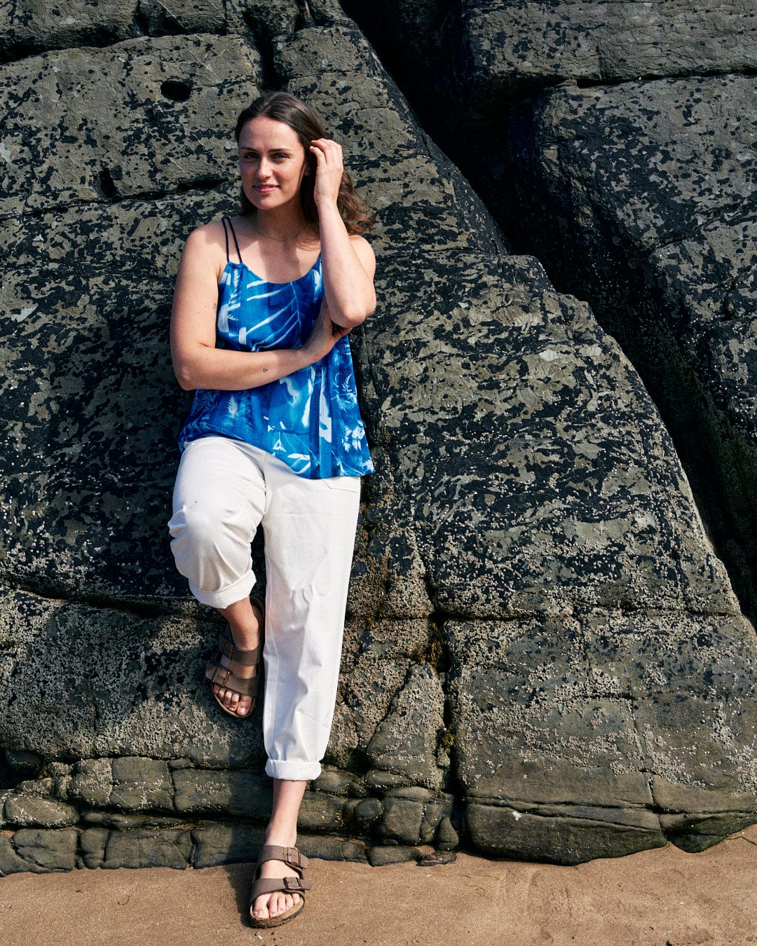 Saltrock Cyanotype - Womens Cami - Blue and white pants stand next to a textured rock formation, with one foot resting on the rock, and her hand touching her hair.