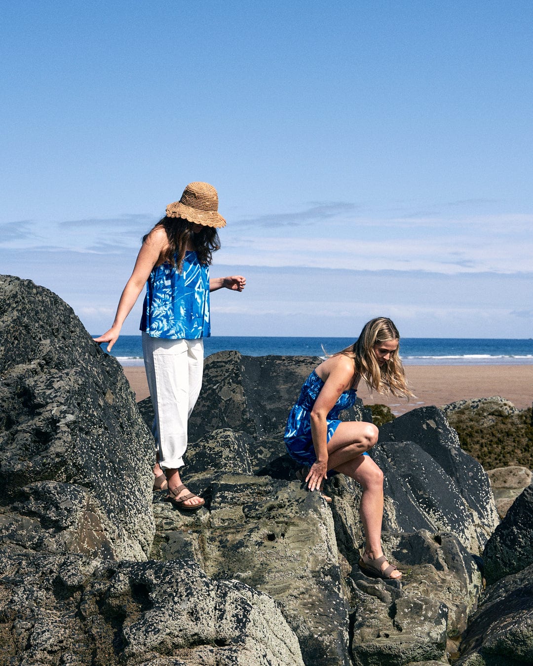 Two women in Cyanotype - Womens Cami - Blue by Saltrock and summer hats, made from lightweight material, climb large rocks on a beach under a clear blue sky. One of them is stepping down while the other stands and looks around.