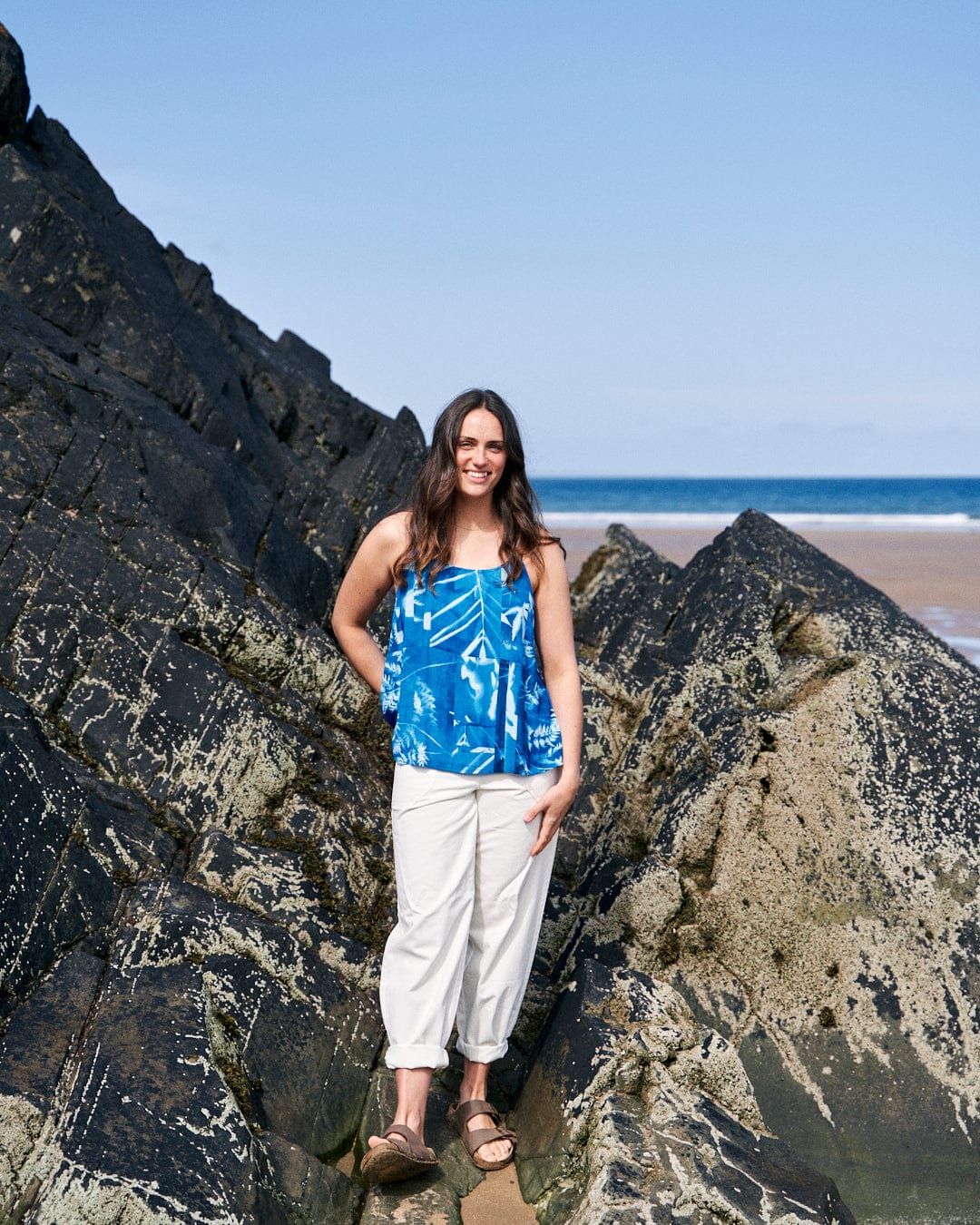 A woman stands on a rocky beach, wearing the Cyanotype - Womens Cami - Blue from Saltrock, white pants, and sandals. The background features a clear sky and a sandy shoreline. The cami's abstract floral print adds a touch of elegance to the serene scene.