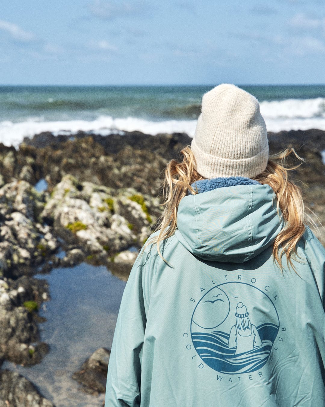 A person wearing the Saltrock Recycled Changing Robe in light green and a beige beanie stands facing the rocky shoreline and ocean waves.