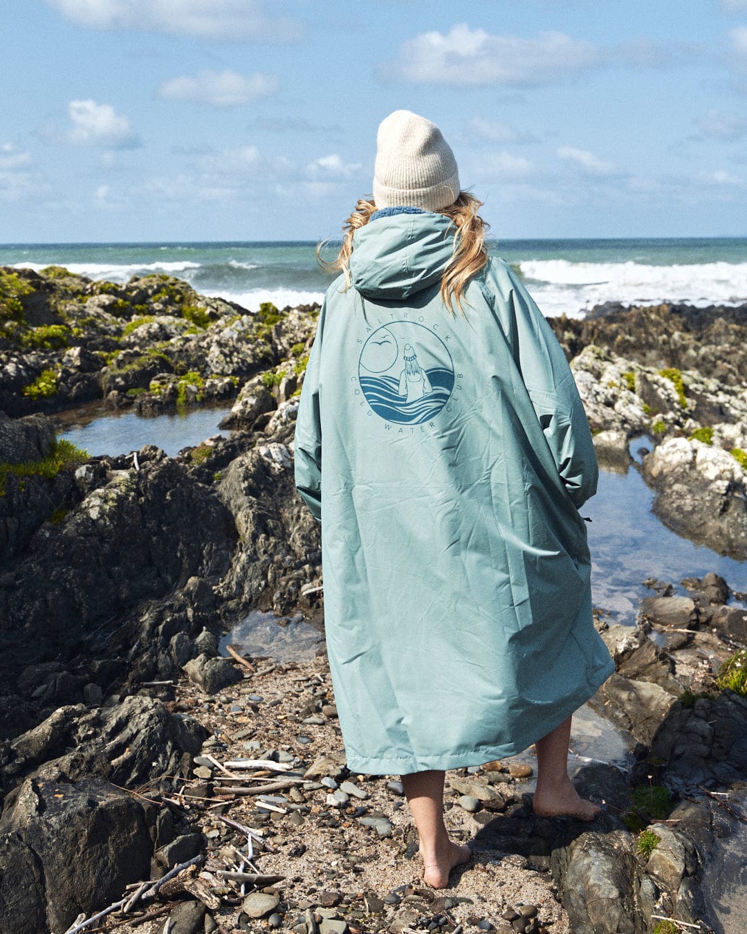 Wearing a Saltrock Recycled Changing Robe in light green and a beige beanie, an individual stands on the rocky shore, gazing out at the ocean.