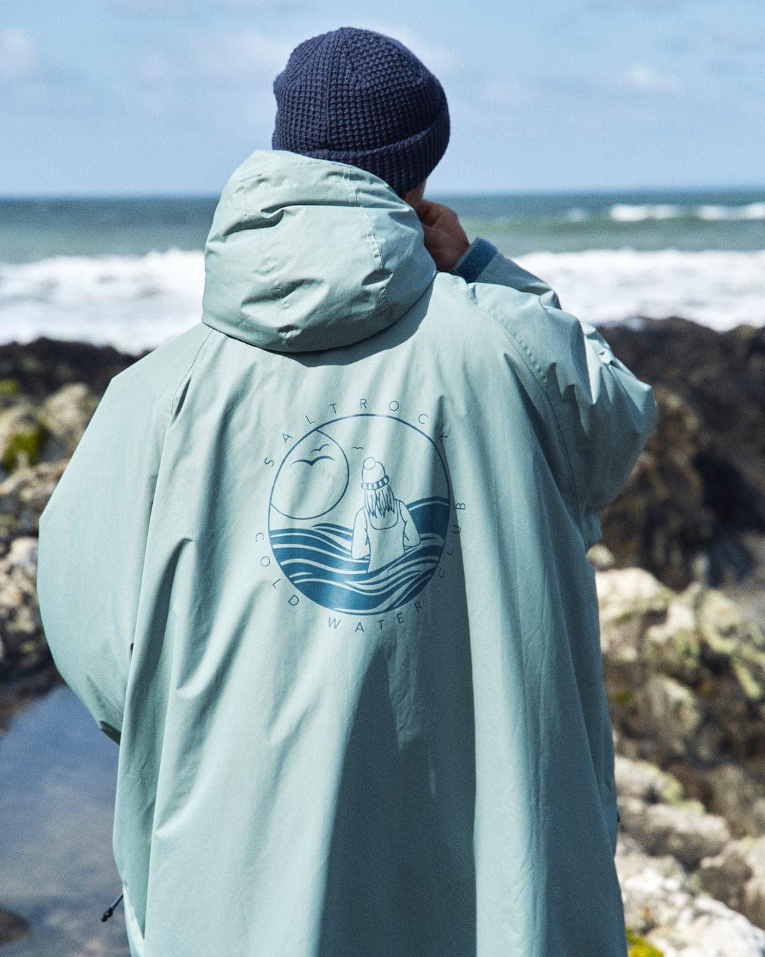 A person in a light green Recycled Changing Robe from Saltrock stands at the rocky shoreline, facing the ocean. The back of the robe features a circular wave and sunset design with text, complementing their knit cap in the cool sea breeze.