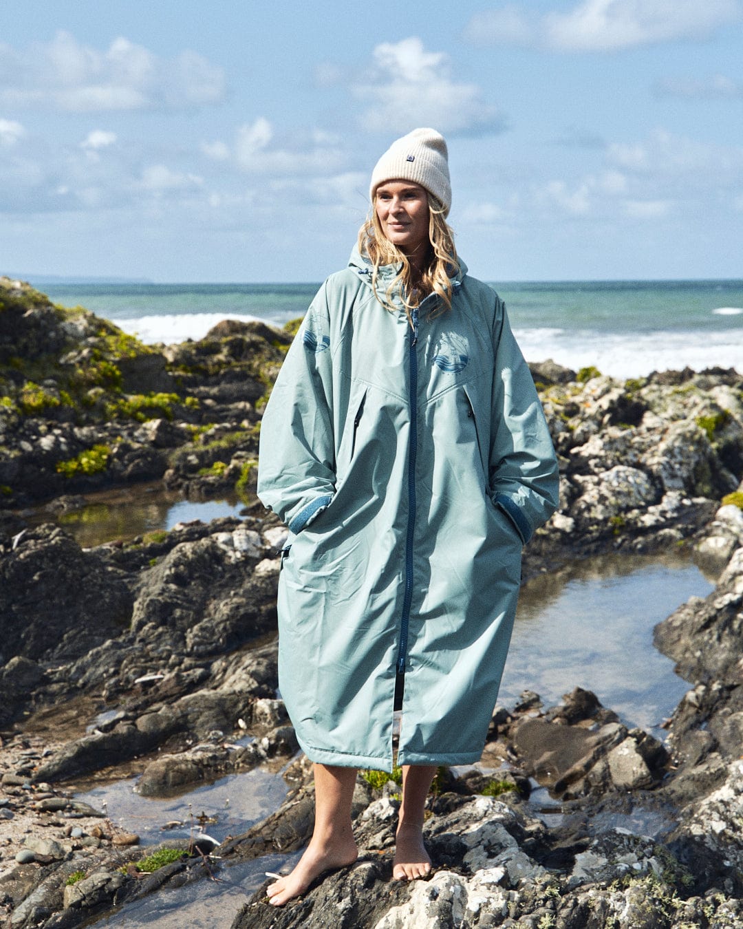 A person in a light green Recycled Changing Robe from Saltrock and a white beanie stands barefoot on the rocky shore, with the ocean and cloudy sky in the background.