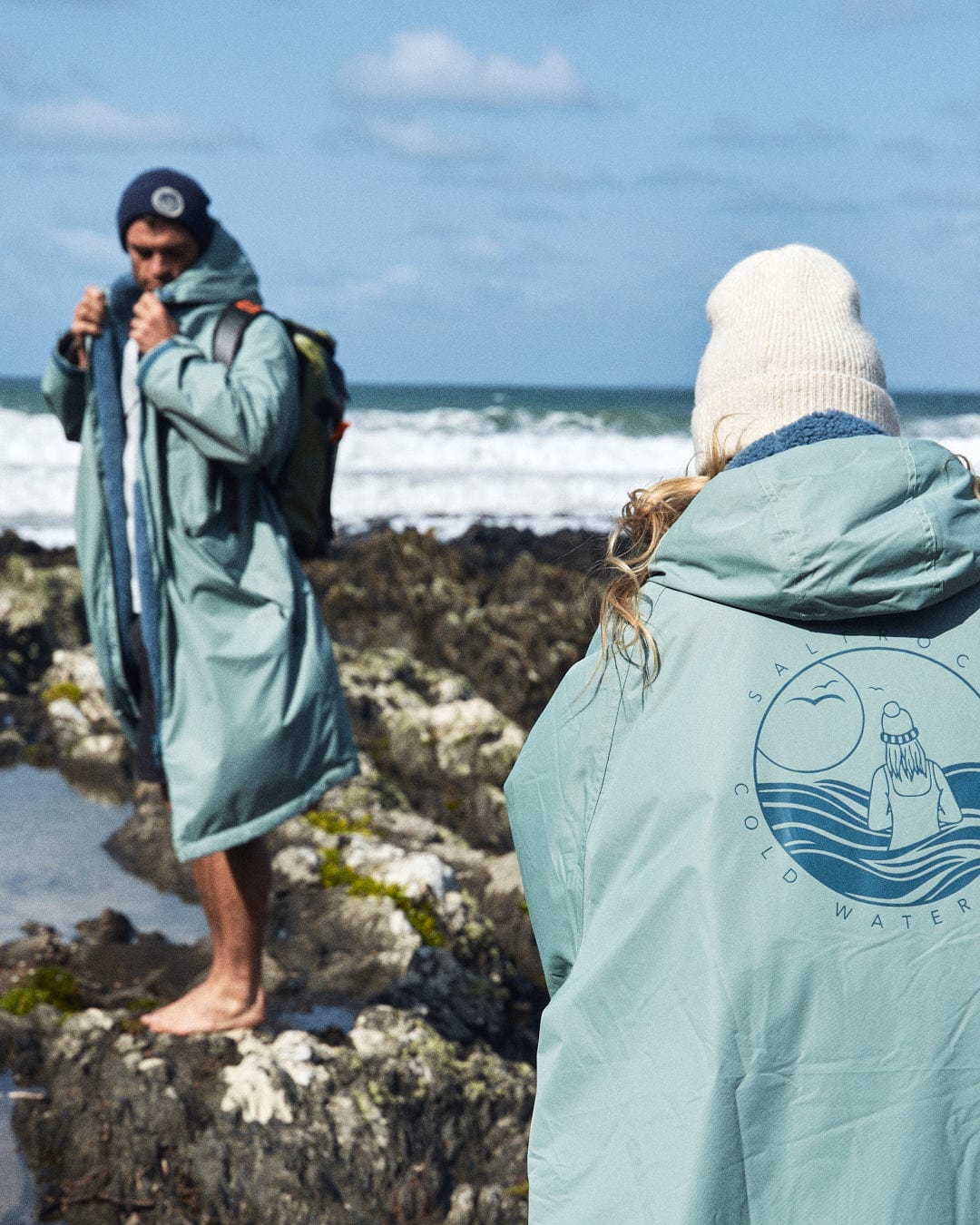 Two individuals wearing Saltrock's Recycled Changing Robe in light green stand on the rocky shore by the ocean. One adjusts a scarf while the other faces away, both clad in robes made from recycled materials, under a cloudy sky.