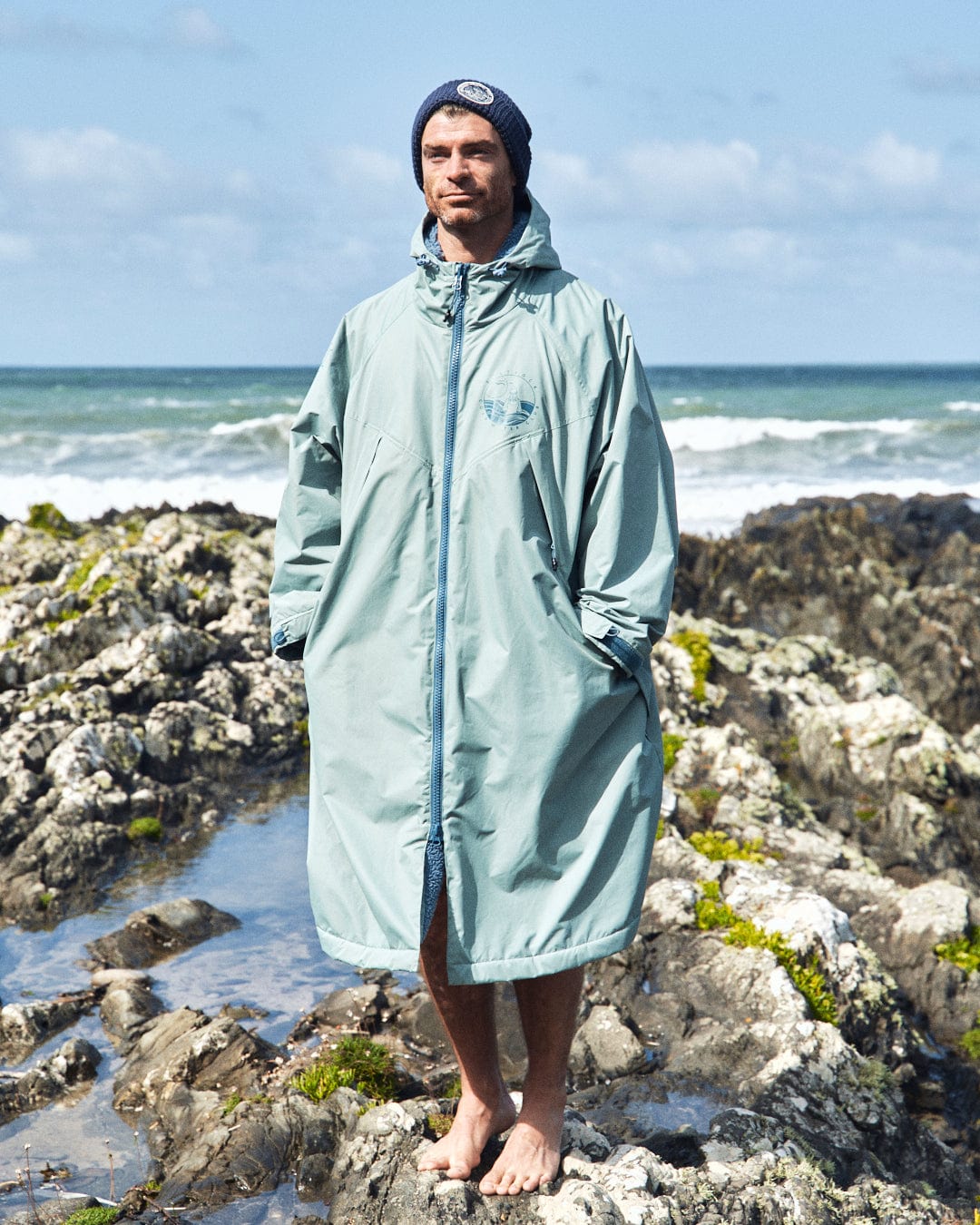 A person stands barefoot on rocky terrain by the ocean, wearing a light green Recycled Changing Robe from Saltrock and a beanie.