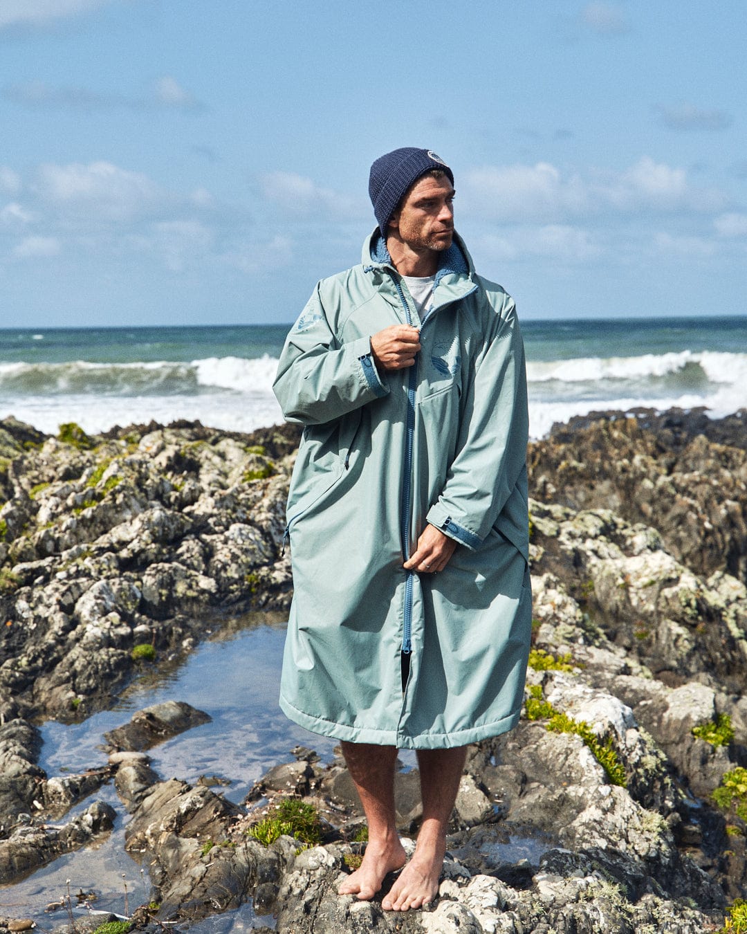 A man stands barefoot on rocky terrain by the sea, wearing a light green Saltrock Recycled Changing Robe and a beanie.