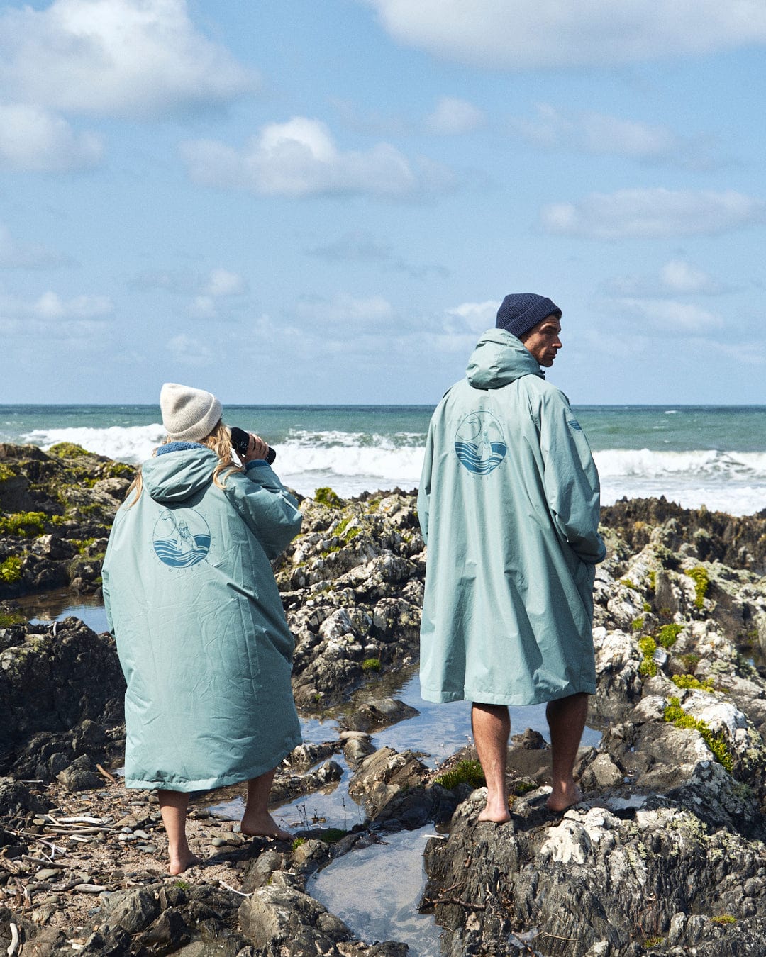 Two individuals wearing the Saltrock Recycled Changing Robe in light green, made from recycled materials, stand on rocky terrain by the ocean, both gazing towards the waves.