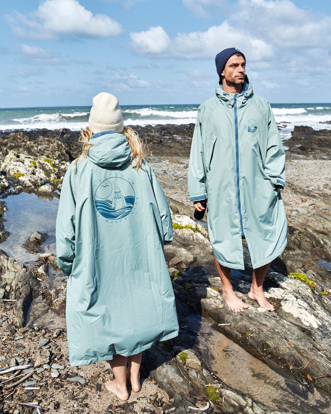 Two individuals wearing Saltrock's Recycled Changing Robes in light green, and beanies stand barefoot on a rocky beach. The ocean and a cloudy sky create a serene backdrop.