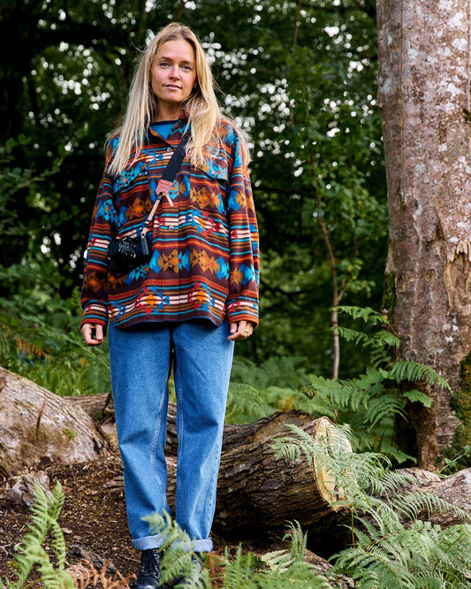 A person with long hair stands in a forest wearing Saltrock's Dallyn Aztec Women's Oversized Fleece Shirt in brown and jeans, with a camera hanging around their neck.