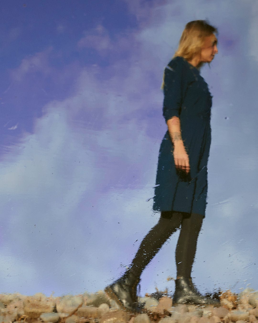 A fashion-conscious woman in a blue dress, the Della - Womens Wrap Top Dress - Dark Blue from Saltrock, gracefully walks through a puddle, showcasing the Saltrock branding on her attire.