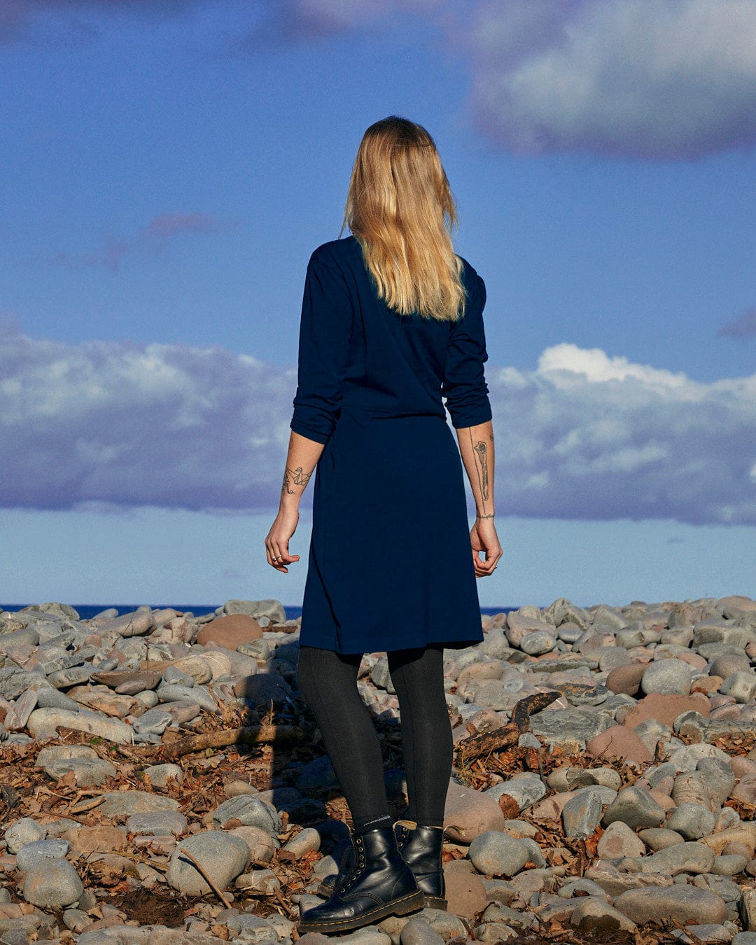 A fashion-conscious woman in a blue dress standing on a rocky beach, wearing the Della - Womens Wrap Top Dress - Dark Blue from Saltrock branding.