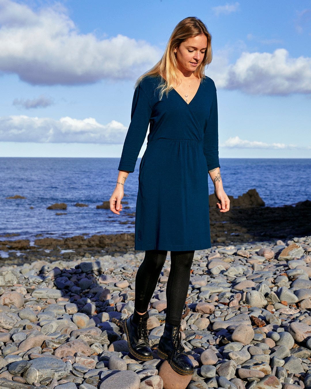 A fashion-conscious woman in a Della - Womens Wrap Top Dress - Dark Blue standing on rocks near the ocean, showcasing the Saltrock branding.