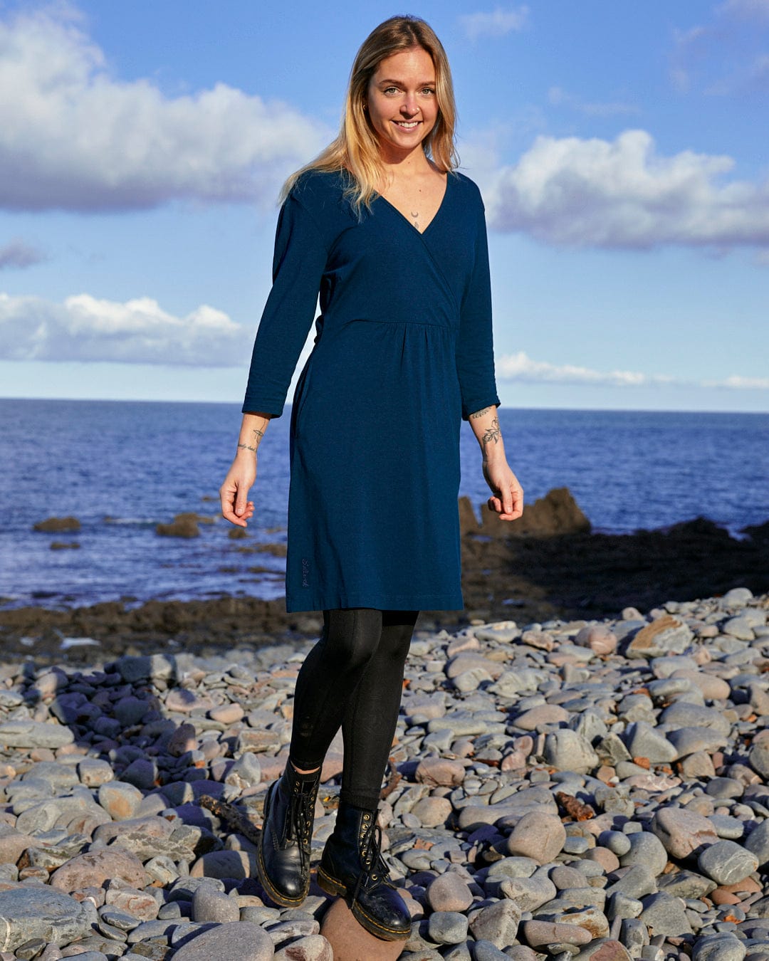 A fashion-conscious woman in a blue dress, the Saltrock Della - Womens Wrap Top Dress - Dark Blue, standing on rocks near the ocean.