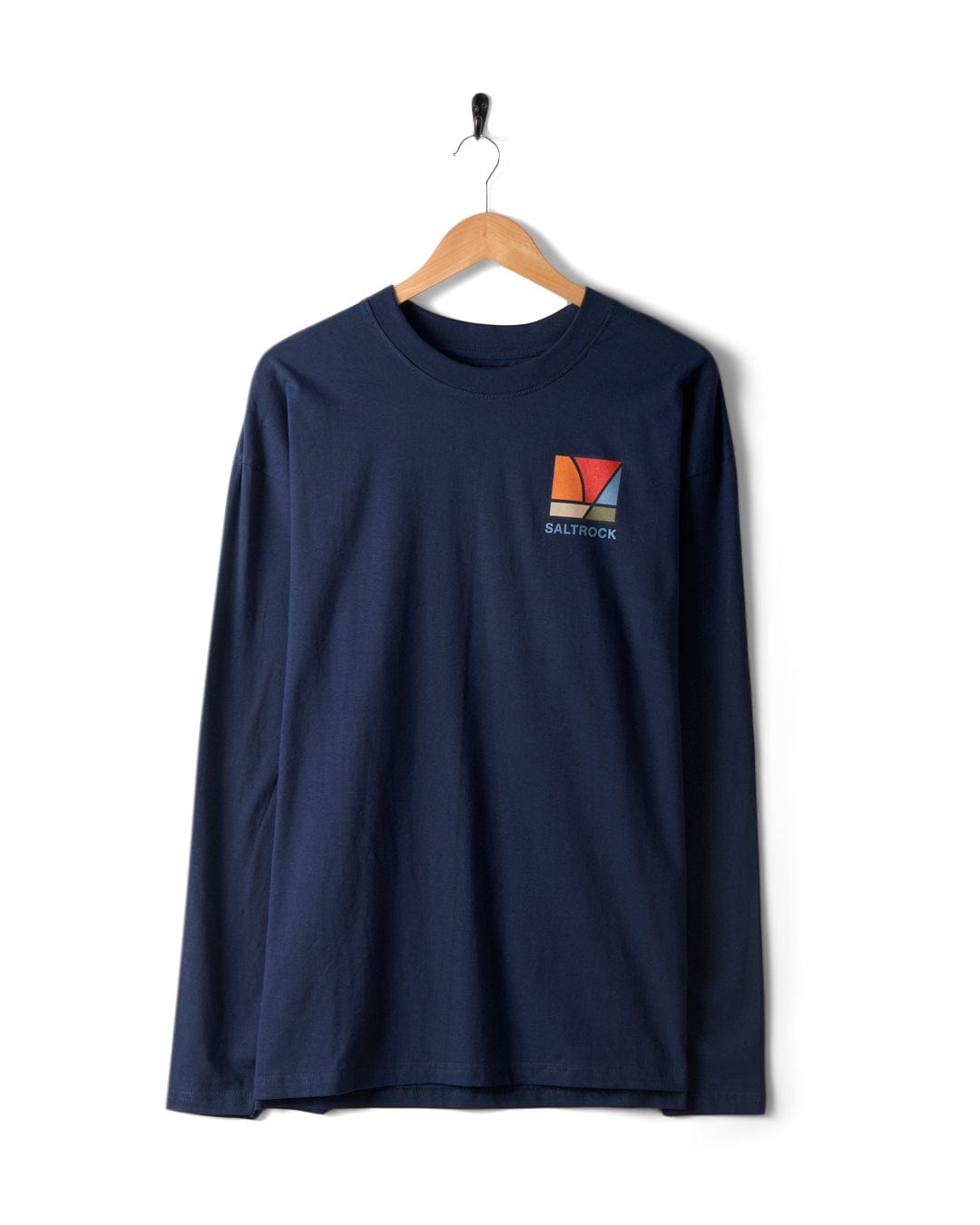 The Saltrock Dimensions Colour 2 Men's Long Sleeve T-Shirt in blue hangs on a wooden hanger against a white background. This 100% cotton shirt features a small geometric logo and "SALTROCK" text on the left chest, enhanced by subtle sunrise prism graphics for an added touch of flair.