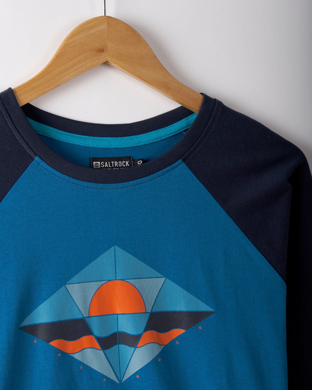 The Dimensions women's long-sleeve t-shirt by Saltrock, in a blue and black raglan style, hangs on a wooden hanger featuring a geometric sunset design on the front. It is crafted from recycled polyester.