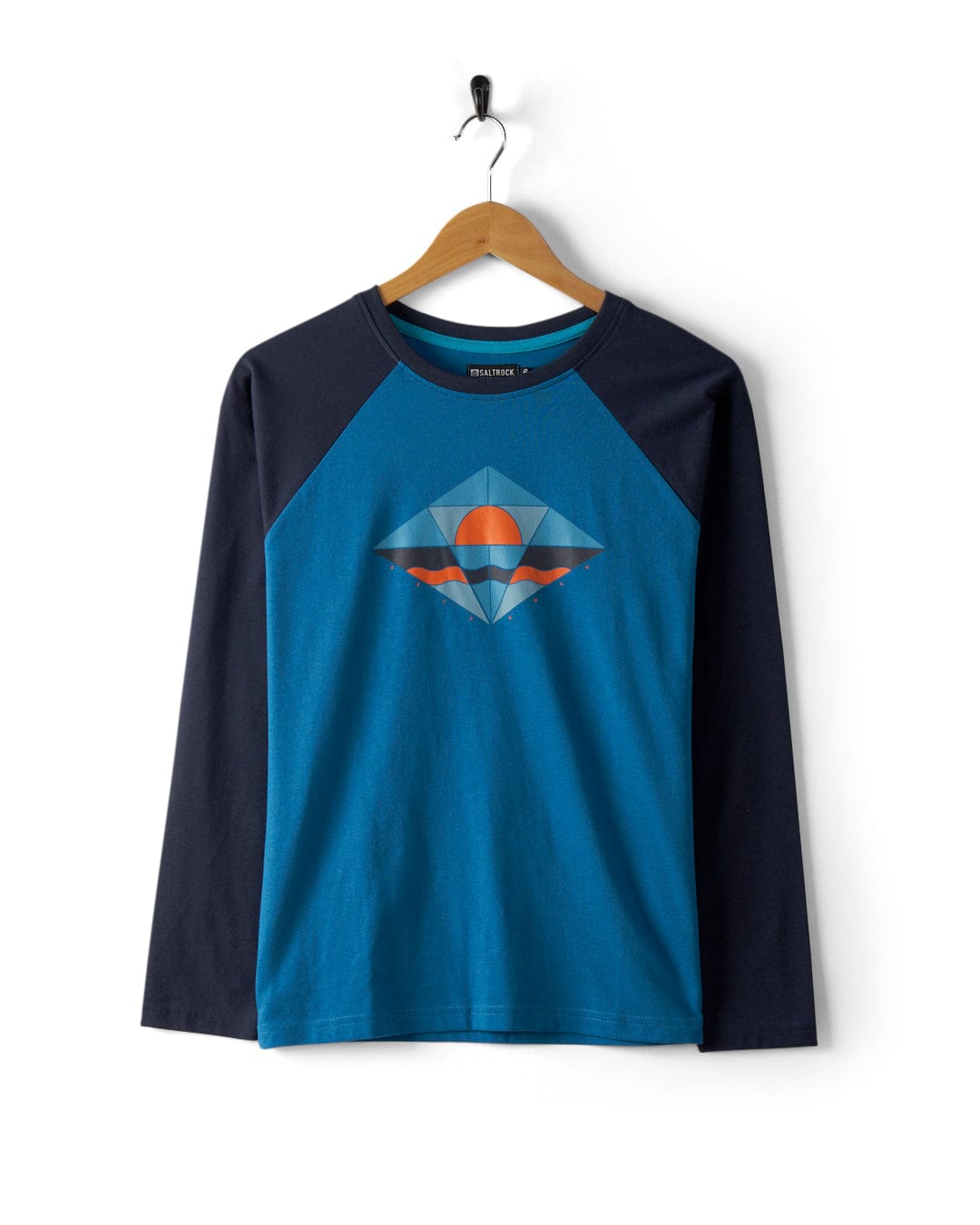 A Saltrock long-sleeve shirt, named the "Dimensions - Womens Long Sleeve T-Shirt - Blue," crafted from recycled polyester, hangs on a wooden hanger. This blue raglan t-shirt features a geometric mountain and sunset graphic on the front, combining style with sustainability.