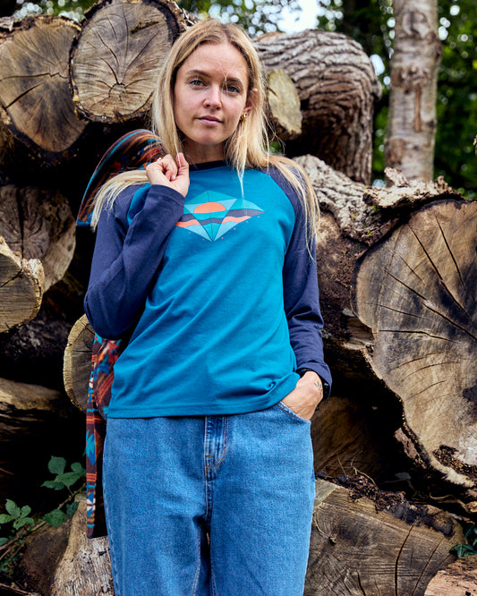 A person wearing the Dimensions women's long-sleeve T-shirt in blue from Saltrock and jeans stands in front of stacked logs outdoors, embracing the spirit of adventure.