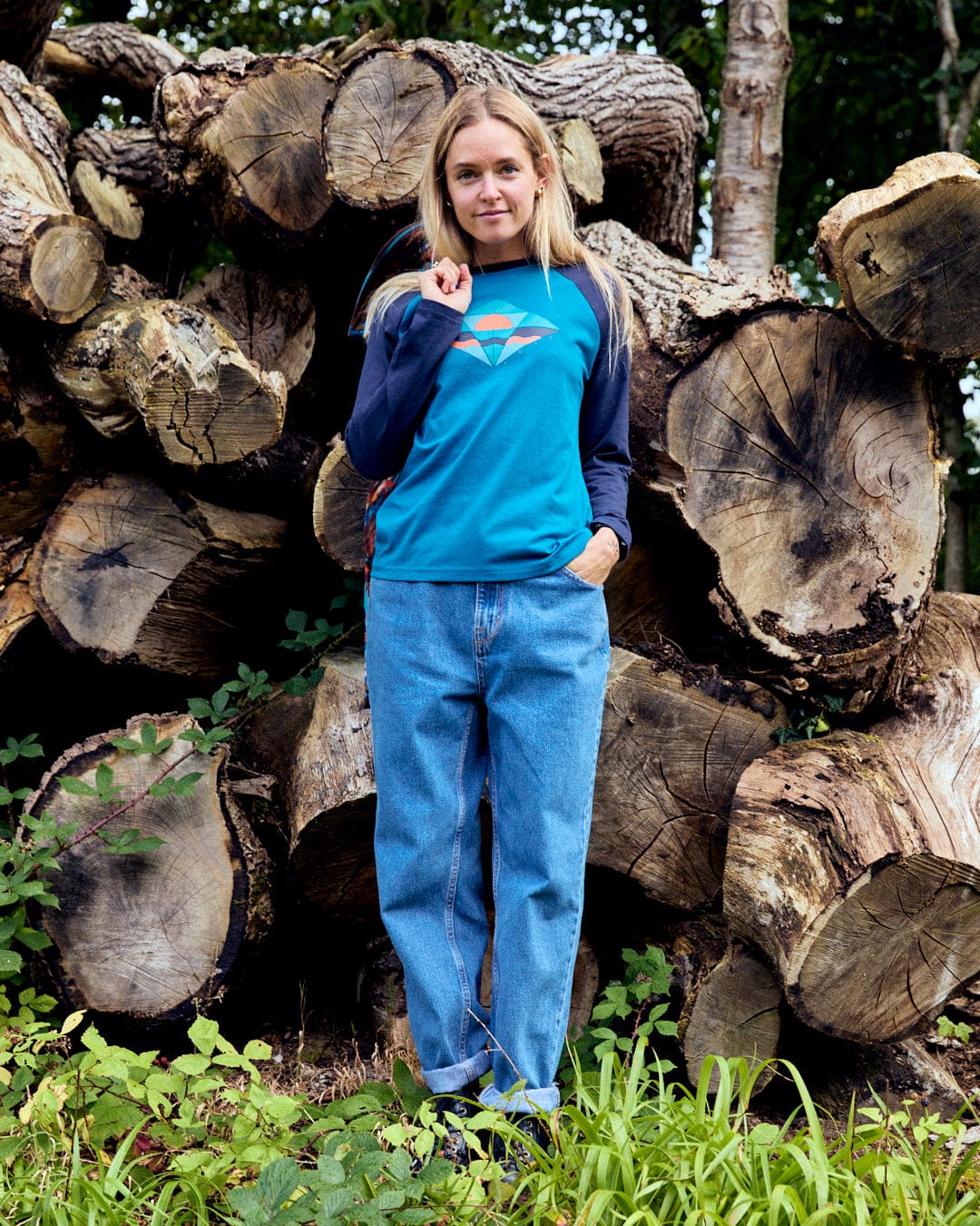 A person wearing a blue Saltrock Dimensions women's long-sleeve T-shirt and jeans stands in front of stacked logs, surrounded by greenery.