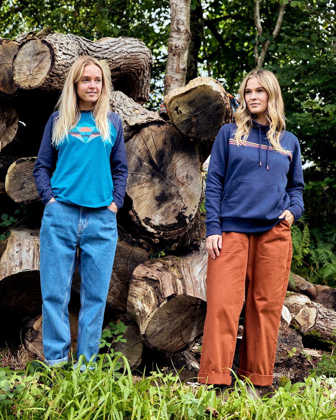 Two women stand in front of stacked logs. One wears Saltrock's Dimensions blue long sleeve t-shirt and jeans, while the other sports a navy hoodie and orange pants. Trees and grass provide a serene backdrop, harmonizing with their casual style.