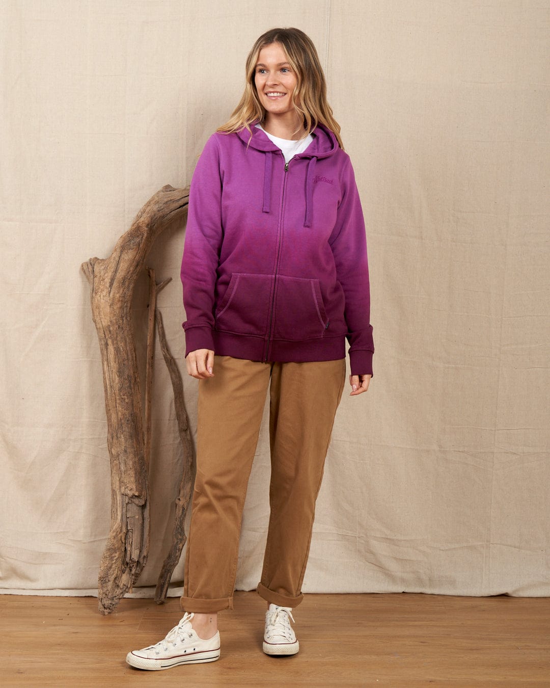 A woman wearing the Saltrock "Dip Dye - Womens Zip Hoodie" in purple and khaki pants stands beside a wooden branch against a beige backdrop, displaying Saltrock branding.