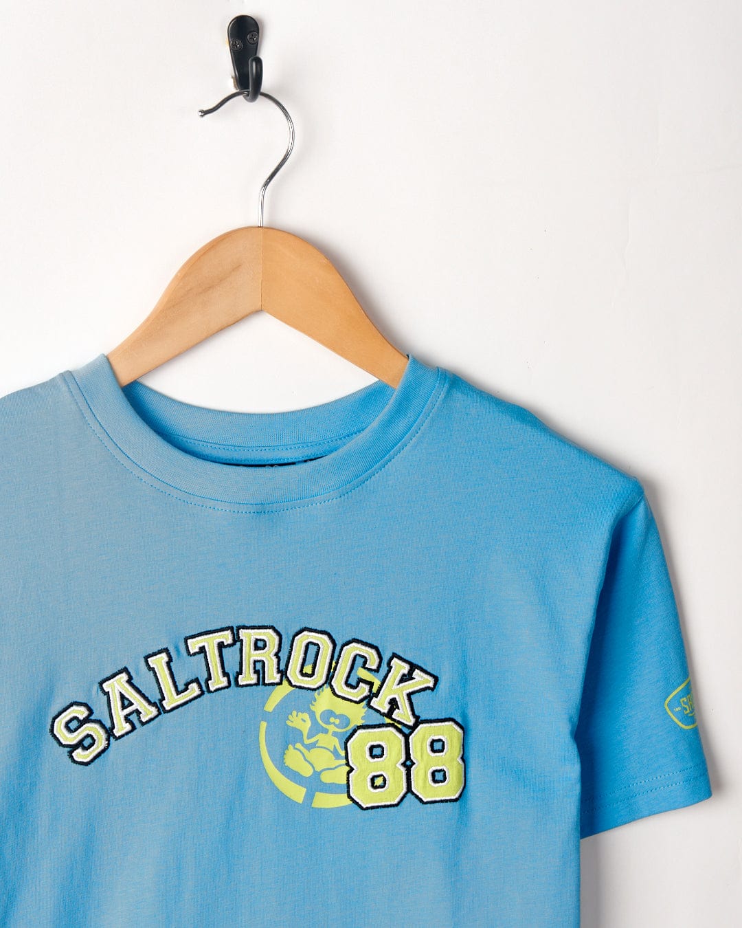 A blue Drop Out - Kids T-Shirt - Blue with "Saltrock 88" and a logo printed on the chest is hanging on a wooden hanger against a white wall. Made from 100% cotton, this stylish tee by Saltrock is not only comfortable but also machine washable for easy care.