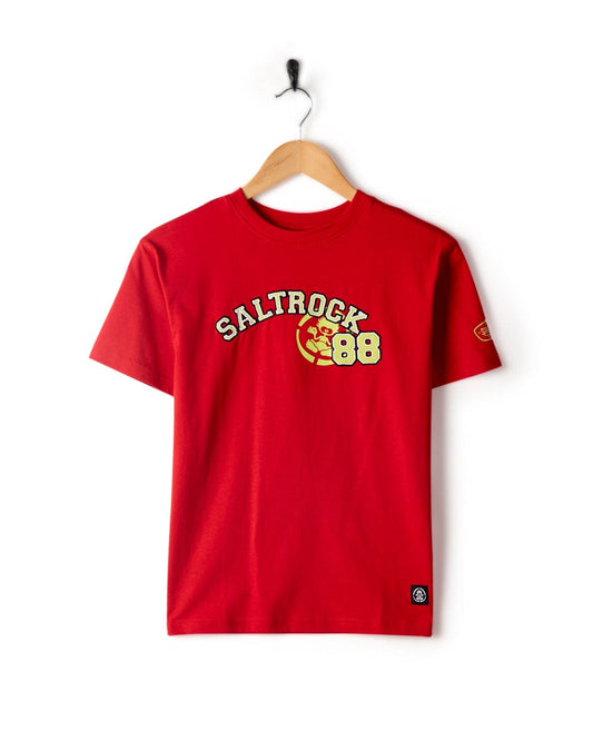 Red 100% cotton T-shirt with the text "Saltrock 88" printed in yellow and white across the chest, featuring a graphic of a wave. Showcased on a wooden hanger against a white background, this tee epitomizes Saltrock branding. This is the Drop Out - Kids T-Shirt - Red by Saltrock.