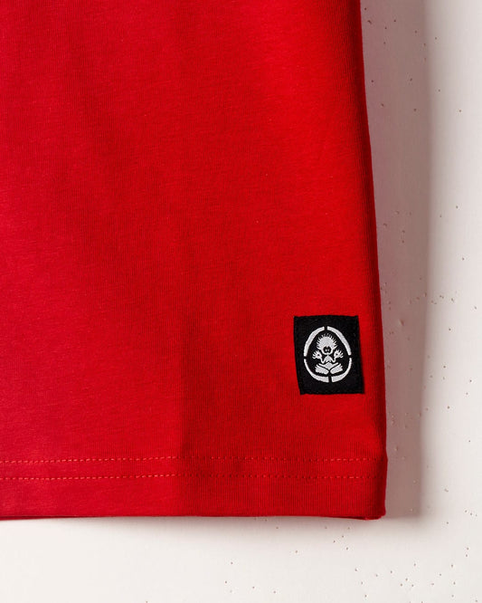 Close-up of the bottom corner of a red t-shirt, the Drop Out - Kids T-Shirt - Red, made from 100% cotton, featuring a small black and white Saltrock branding tag with an image of a skull and two crossed flowers.