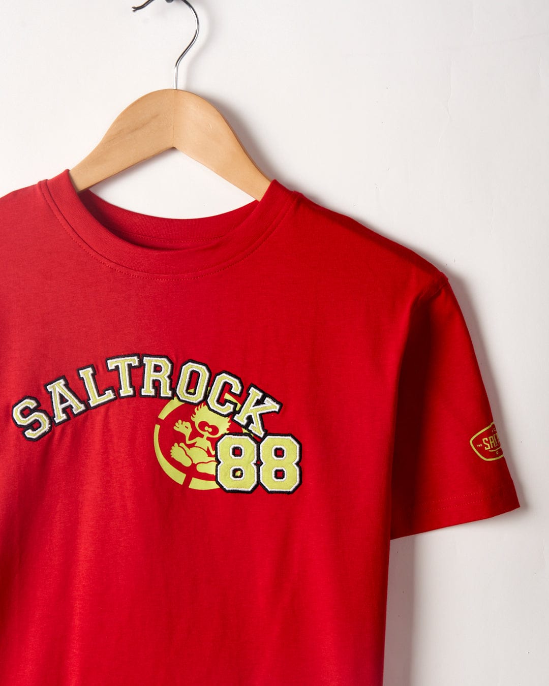 A red Drop Out - Kids T-Shirt - Red, boasting Saltrock branding with a surfer and the number 88 on the front, hangs on a wooden hanger against a white wall. Crafted from 100% cotton, it perfectly combines comfort and style.