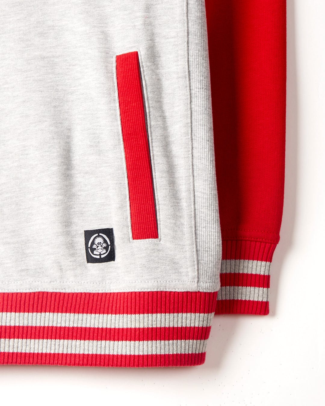 Close-up view of a grey sweatshirt with red raglan sleeves, ribbed red cuffs and waistband featuring white stripes, and a small Saltrock branding patch near the pocket. This is the Saltrock Drop Out - Kids Sweat Shirt - Grey/Red.