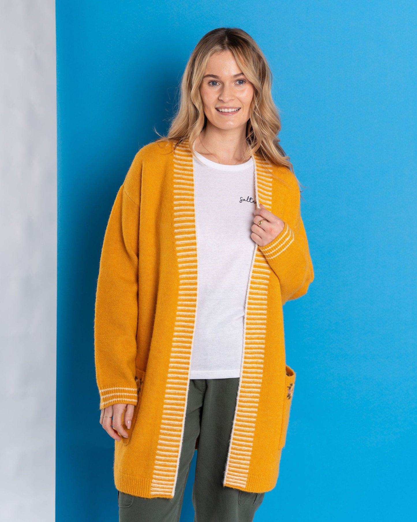 A woman in the Saltrock Coraline Women's Longline Knitted Cardigan in yellow and a white shirt smiles against a blue background.