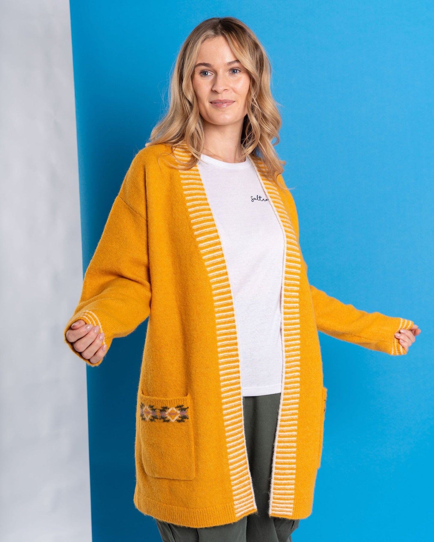 A person wearing a Saltrock "Coraline - Women's Longline Knitted Cardigan - Yellow" over a white shirt stands against a blue background.