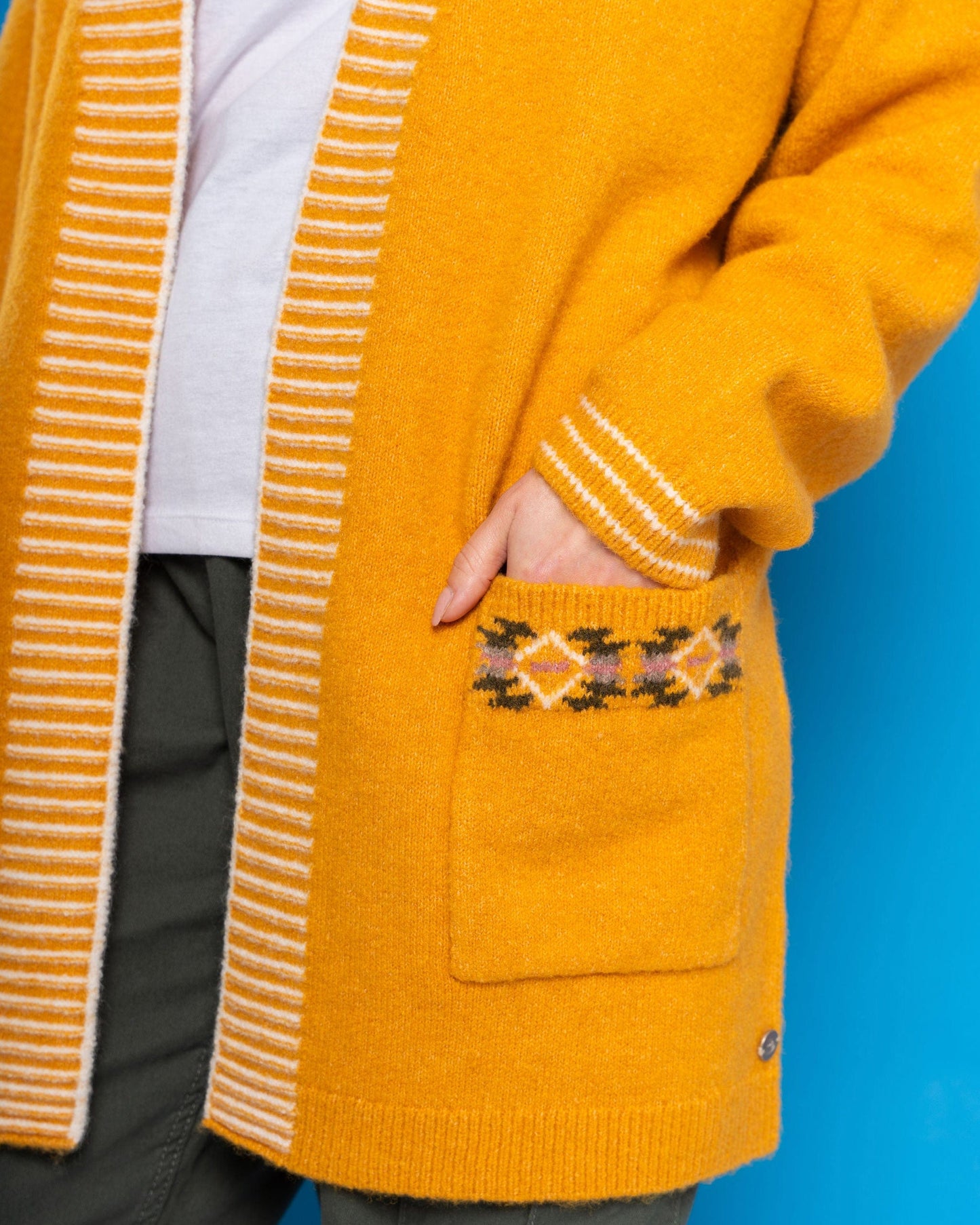 A person dons Saltrock's Coraline - Women's Longline Knitted Cardigan in yellow, featuring an open-front style with decorative pockets, paired with green pants against a blue backdrop.