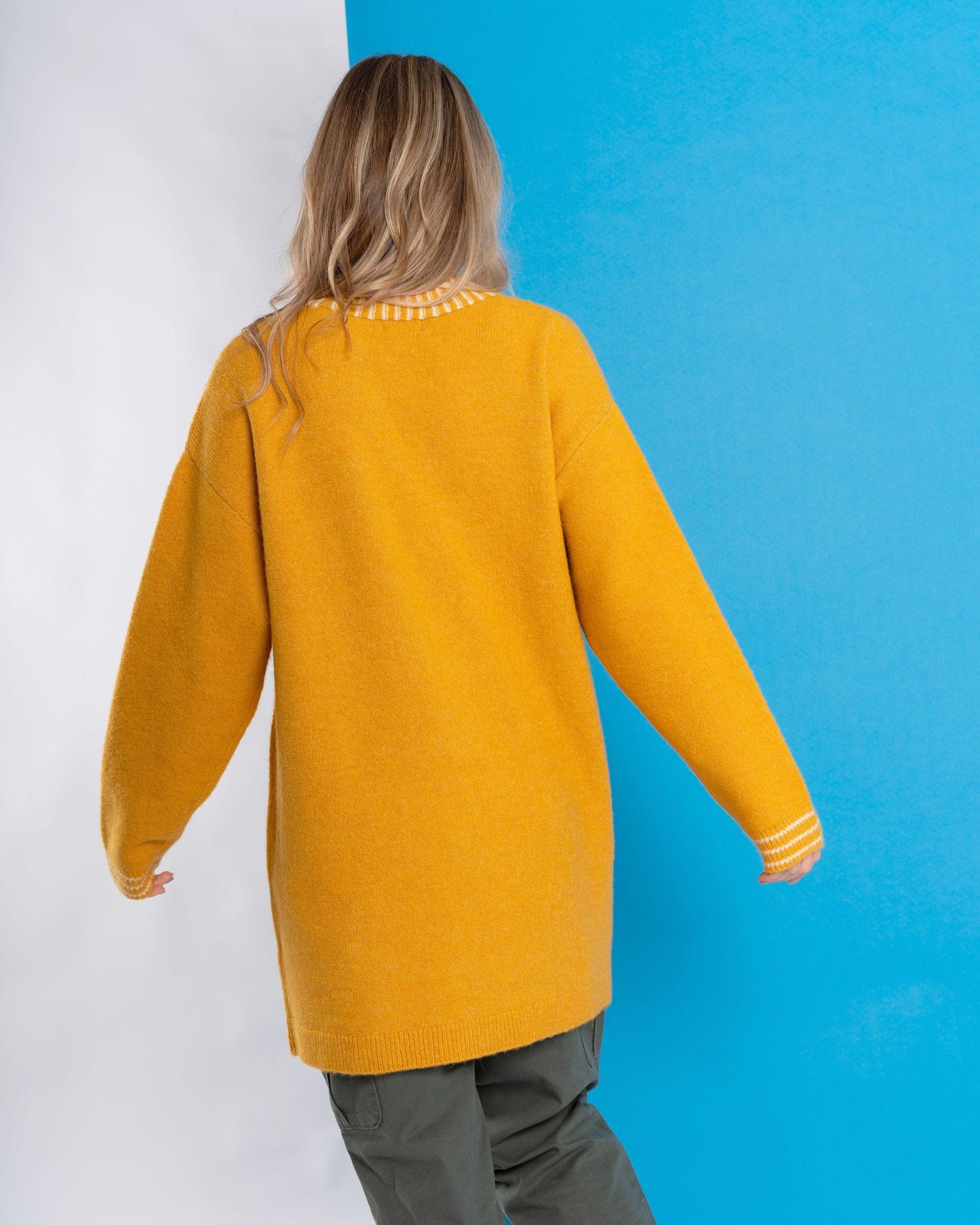 Wearing a Coraline Women's Longline Knitted Cardigan in Yellow by Saltrock, a person with long hair stands against a blue and white backdrop, adding intrigue to the subtle Aztec-patterned scene.