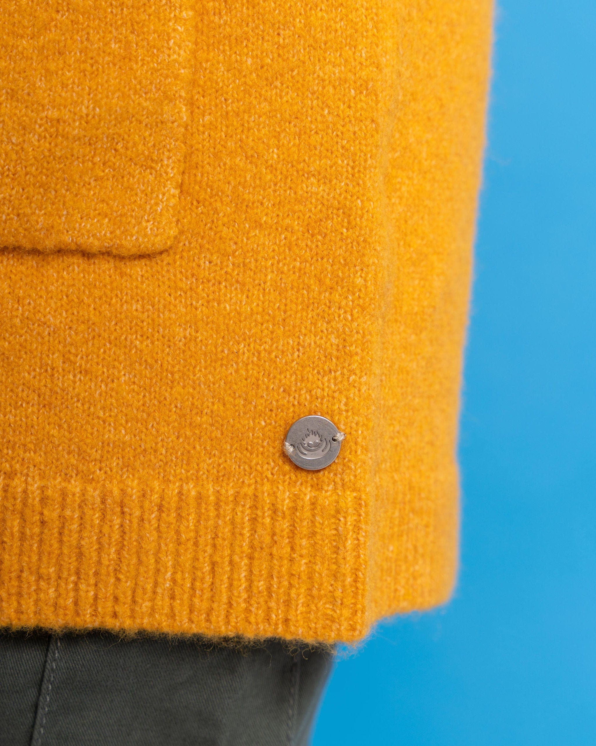 Close-up of the Coraline by Saltrock, a mustard yellow longline knitted cardigan with an open-front style and a metal circular button near the hem, set against a blue background.