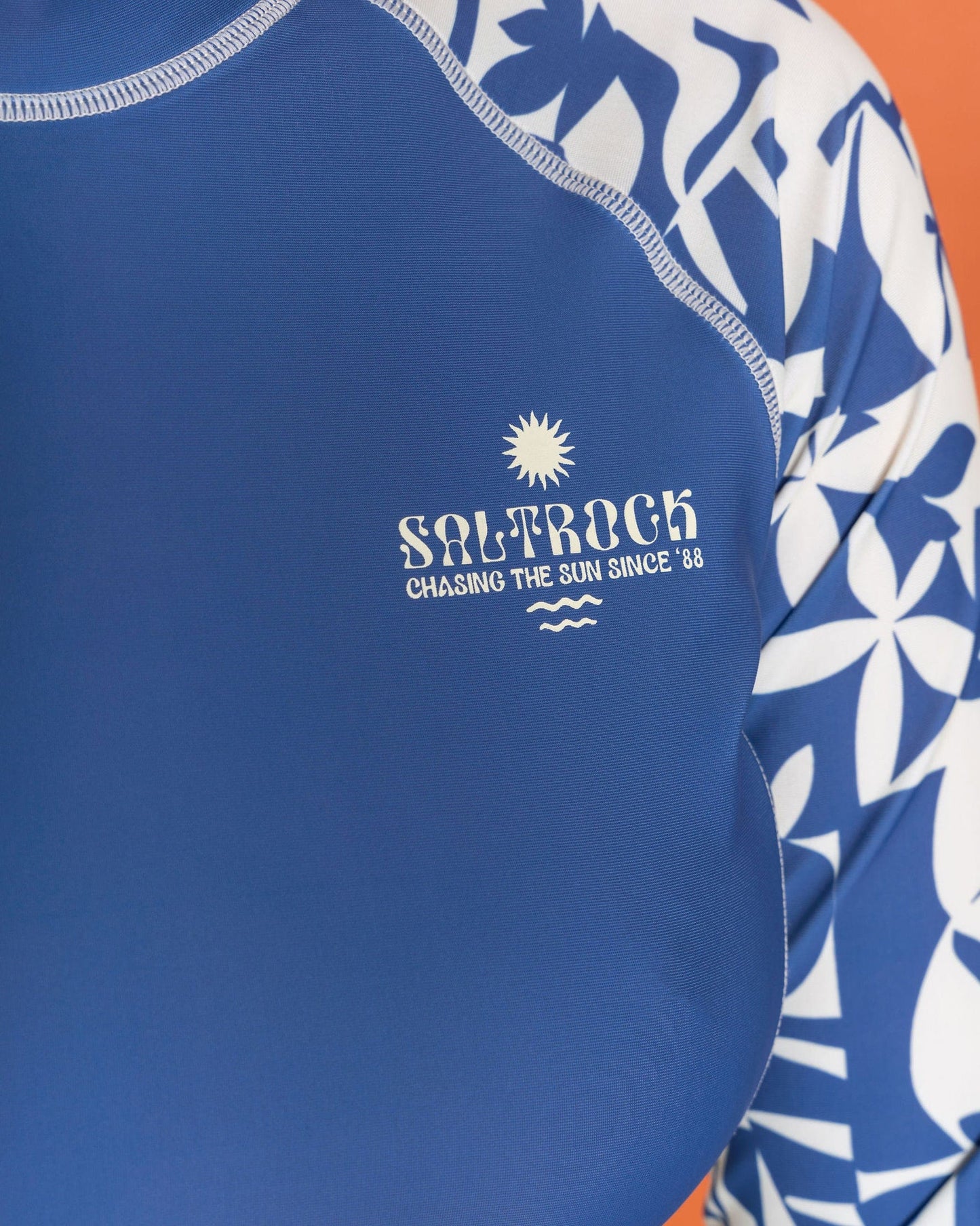 Close-up of the Saltrock "Balance" women's UPF 50+ rashvest in blue adorned with a vibrant floral print; made from recycled polyester, it features the text "Saltrock Chasing the Sun Since '88.