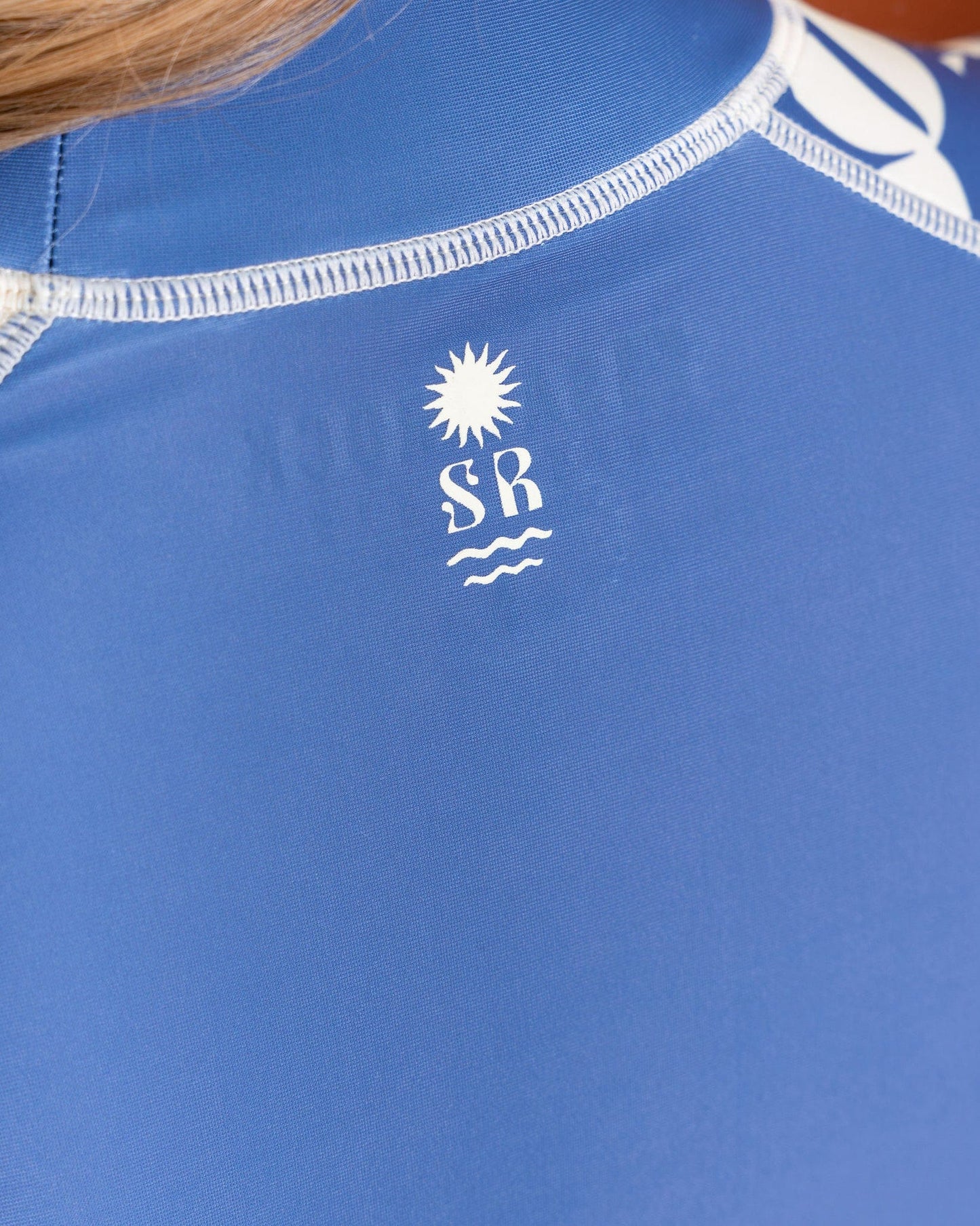 Close-up of the Balance Women's UPF 50+ Rashvest in blue by Saltrock, made from recycled polyester with white stitching. The upper back sports a sun over "SR" and wavy lines logo.