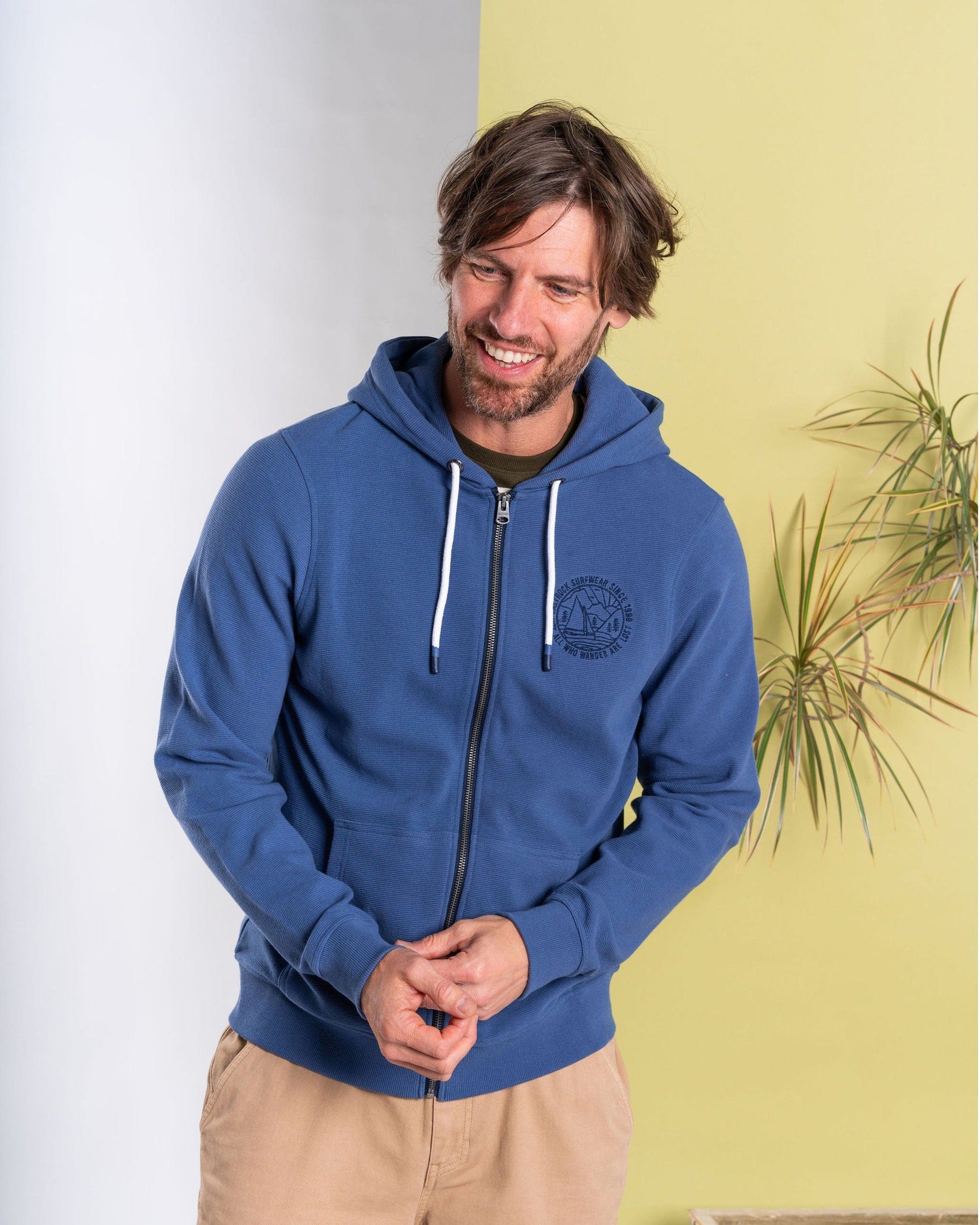 A man wearing the Saltrock Hanns Men's Zip Hoodie in blue stands smiling against a light yellow and white background, hands together, with a plant visible to his right.