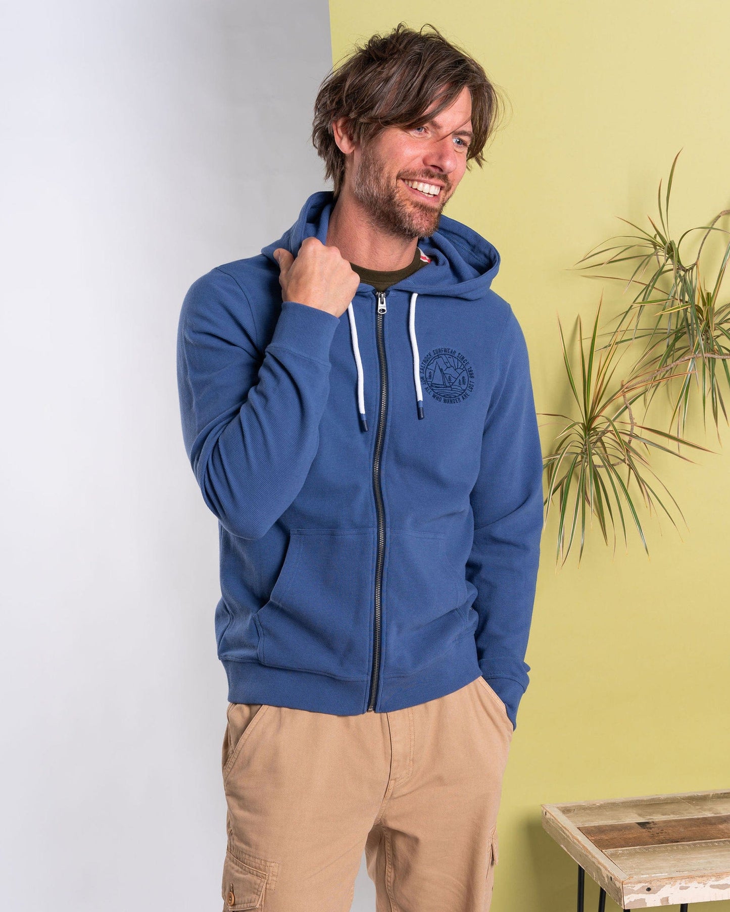 A man in the blue Hanns Saltrock Zip Hoodie, with ribbed texture, stands smiling against a yellow and white backdrop, one hand on his hood. He wears beige pants as a plant and table are partially visible.