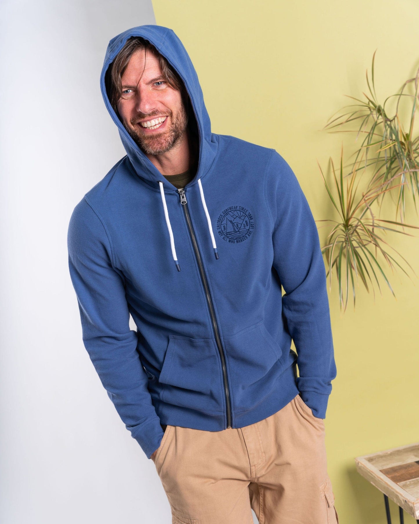 A person in the blue Saltrock "Hanns - Men's Zip Hoodie" smiles while wearing beige pants, standing in front of a pale yellow wall accented by a nearby plant.