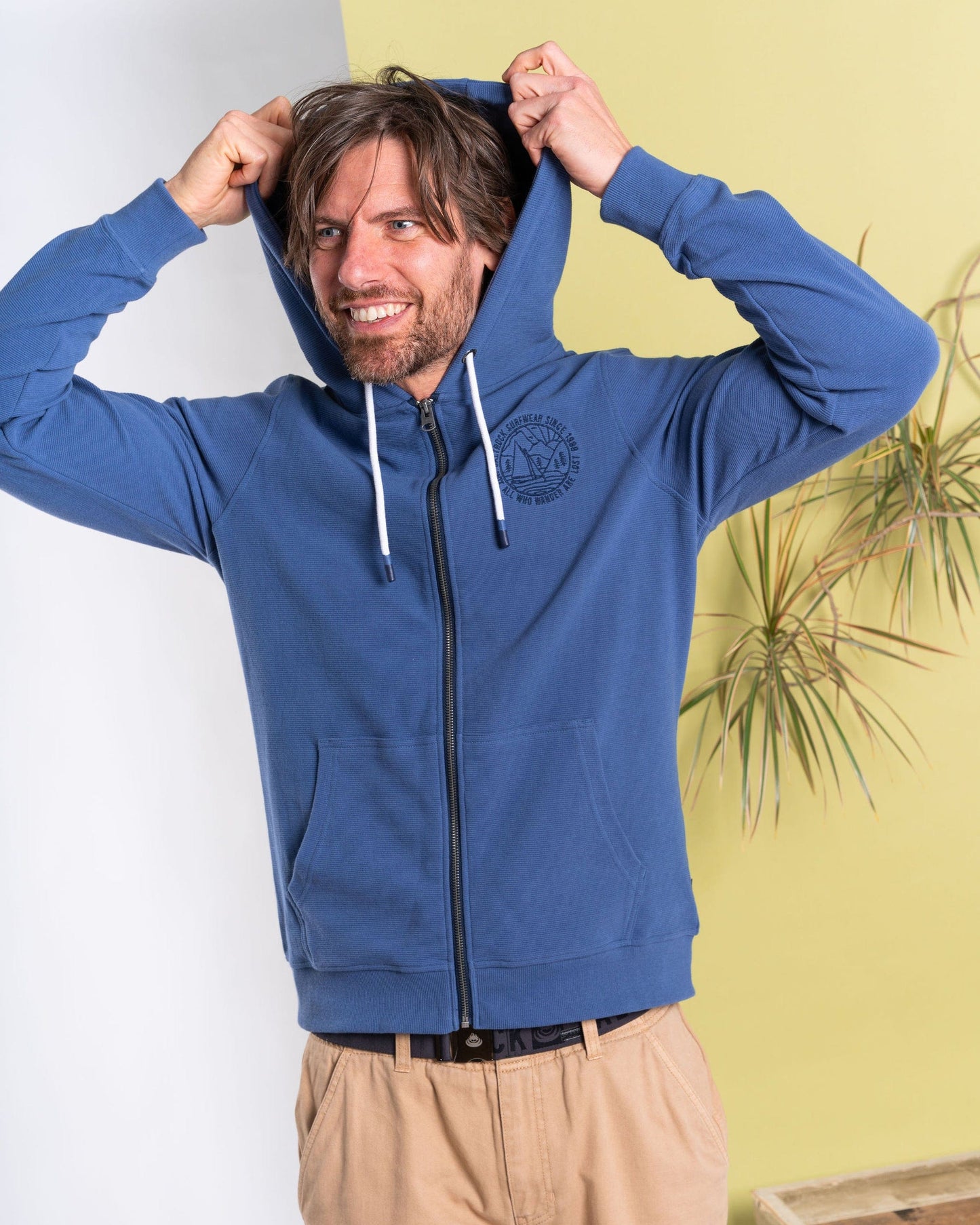 A smiling man poses with the hood on his head, wearing the Hanns - Men's Zip Hoodie in blue by Saltrock. Crafted from cozy 100% cotton, his attire features a ribbed texture and stands out against a yellow wall accented by a potted plant.